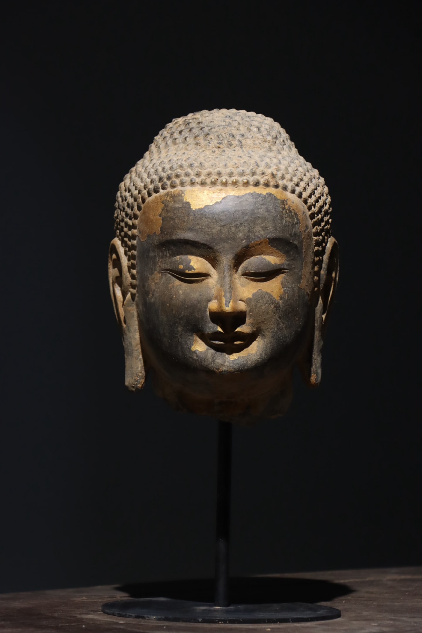 TANG DYNASTY GILT-BRONZE STONE HEAD OF BUDDHA: Total H: 16.14in (Total H: 41.0cm); Net H: 9.84in (Net H: 25.0cm)This Tang Dynasty stone Buddha head features fine snail-shell curls, a serene face with a gentle smile, and traces of gilding. Carved w