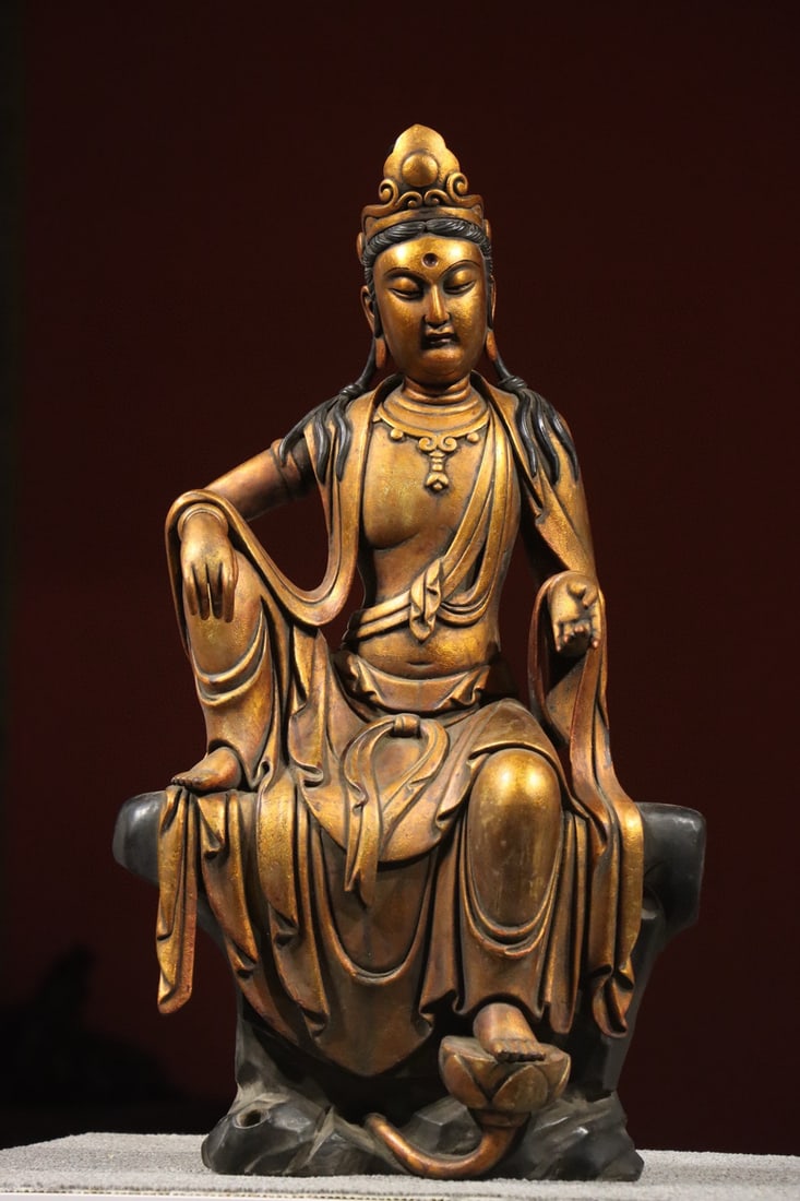 A GILT-LACQUERED WOOD FIGURE OF GUANYIN IN ROYAL EASE, MING DYNASTY, 14TH–17TH CENTURY: Width x Height: 14.57in x 25.59in (37.0cm x 65.0cm)Carved from wood and covered in gold lacquer, this Ming Guanyin sits in royal ease with a serene expression. Its ornate crown, flowing robes, and gil