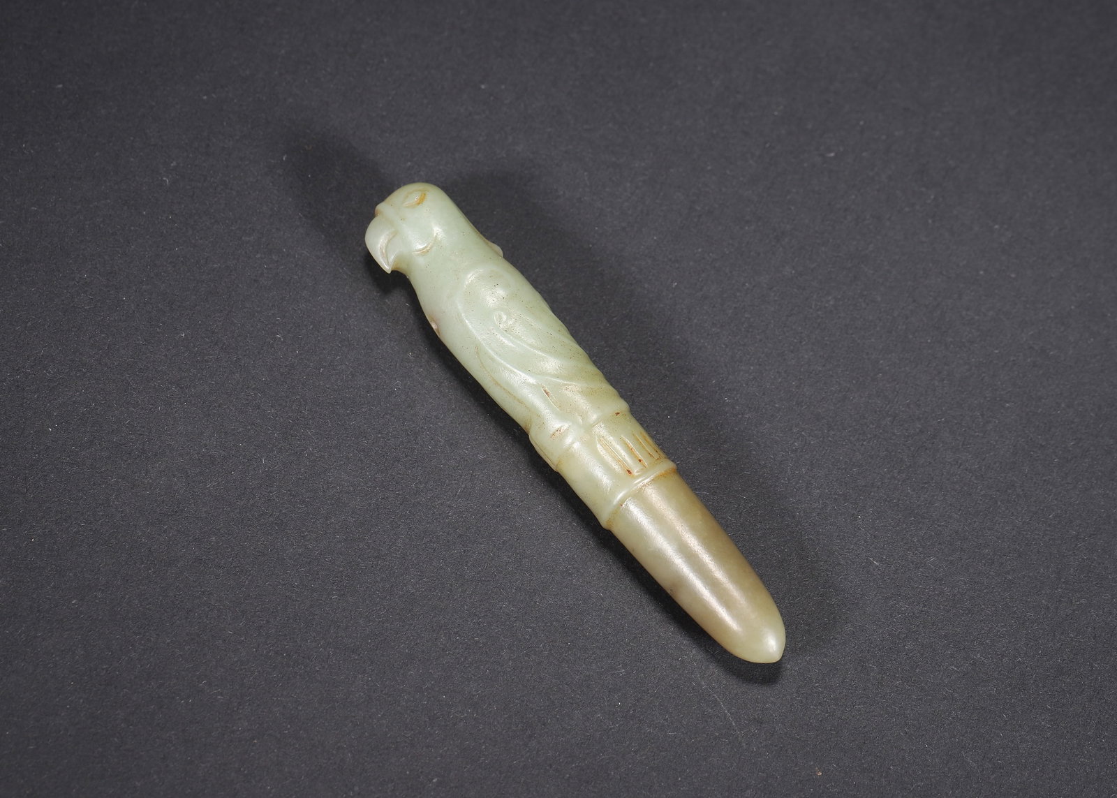 MING DYNASTY PALE GREEN JADE BIRD-HEAD HAIRPIN: 2.91" x 0.47" (7.4cm x 1.2cm) 0.037lb (17g)A fine Ming Dynasty pale green jade hairpin, carved as a bird head with incised feather patterns. Featuring natural russet inclusions and a piercing for cord