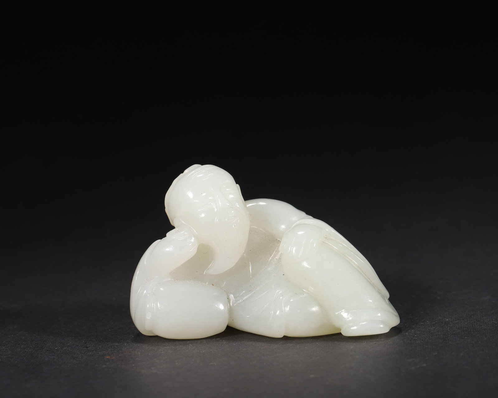 A FINE HETIAN WHITE JADE CARVING OF A RECLINING SCHOLAR, MID-QING DYNASTY: 1.97in x 1.38in (5.0cm x 3.5cm), 0.157lb (71.0g)This well-carved Mid-Qing Hetian white jade depicts a scholar reclining against a wine jar, with serene expression and flowing robes. Its lifelike form 