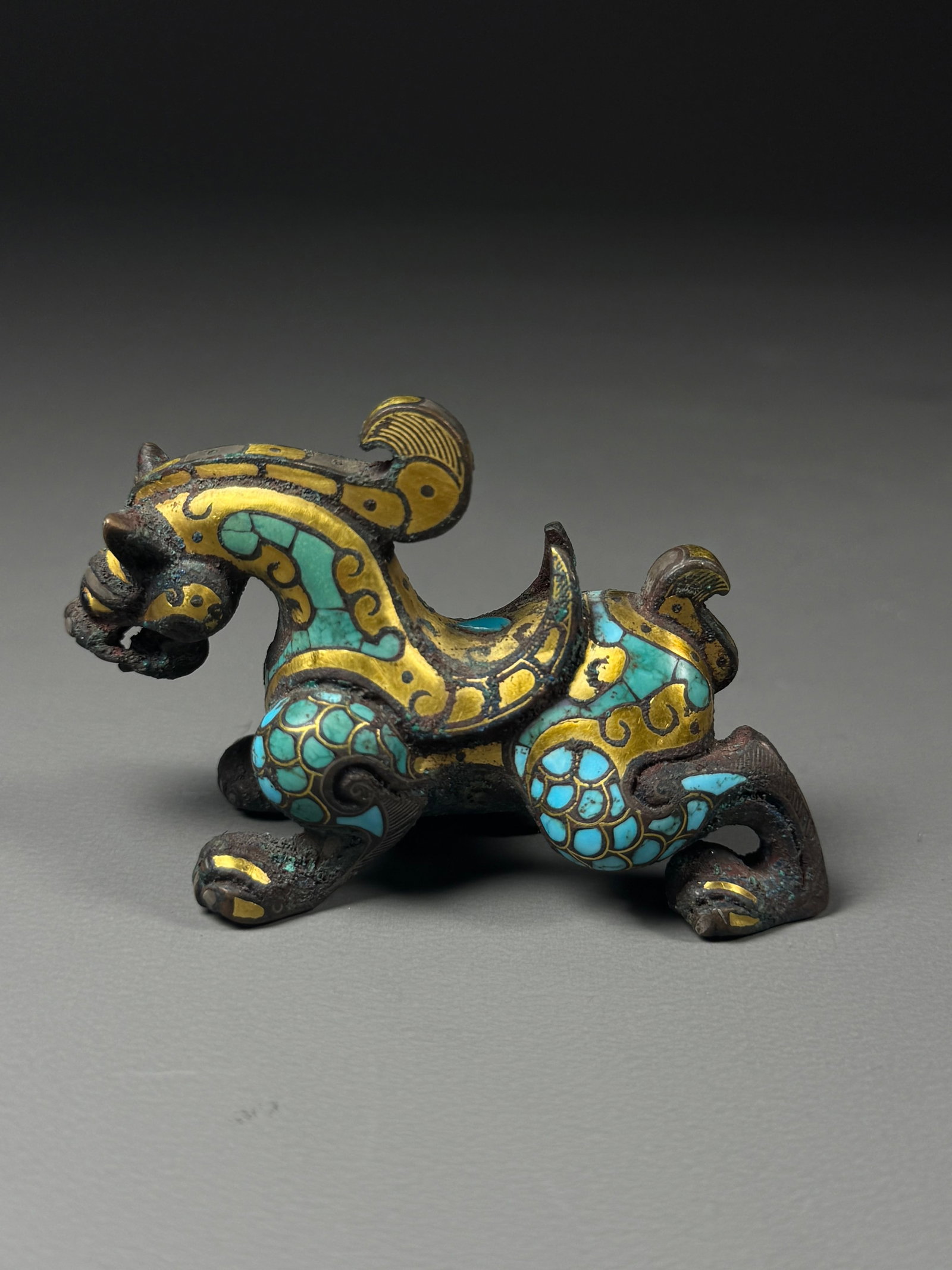 WARRING STATES PERIOD, BRONZE PAPERWEIGHT INLAID WITH GOLD AND TURQUOISE: Warring States Bronze Auspicious Beast Weight Inlaid with Gold & Turquoise. Cast in bronze, this mat weight takes the form of a crouching, backward-glancing mythical beast. Fine gold wire outlines scr