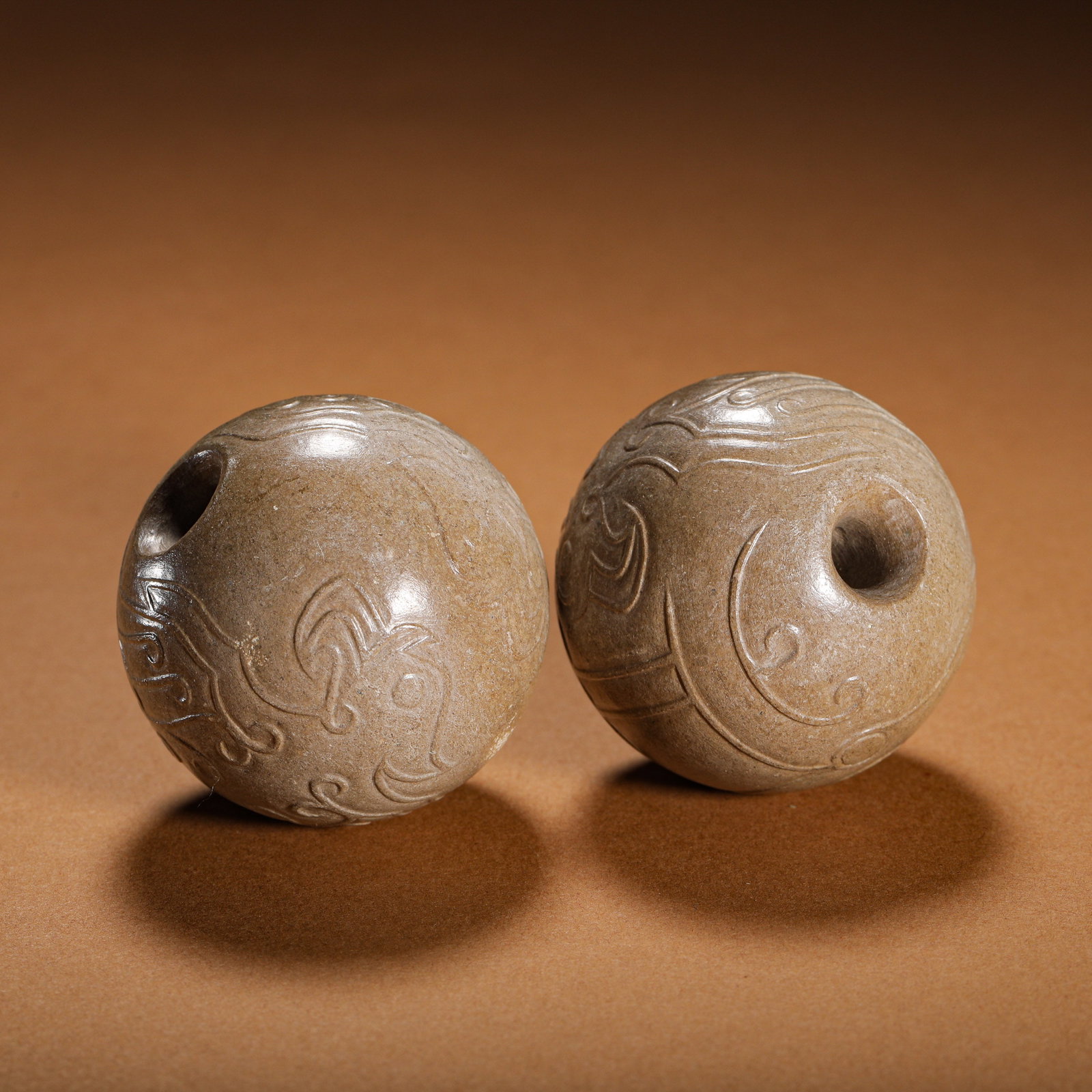 A PAIR OF HAN DYNASTY PHOENIX-PATTERNED JADE BEADS: Pair of spherical jade beads with phoenix scroll carving, Han Dynasty. The round jade beads are fully covered with grey-brown burial patina, each pierced with a straight through-hole. The surface is f