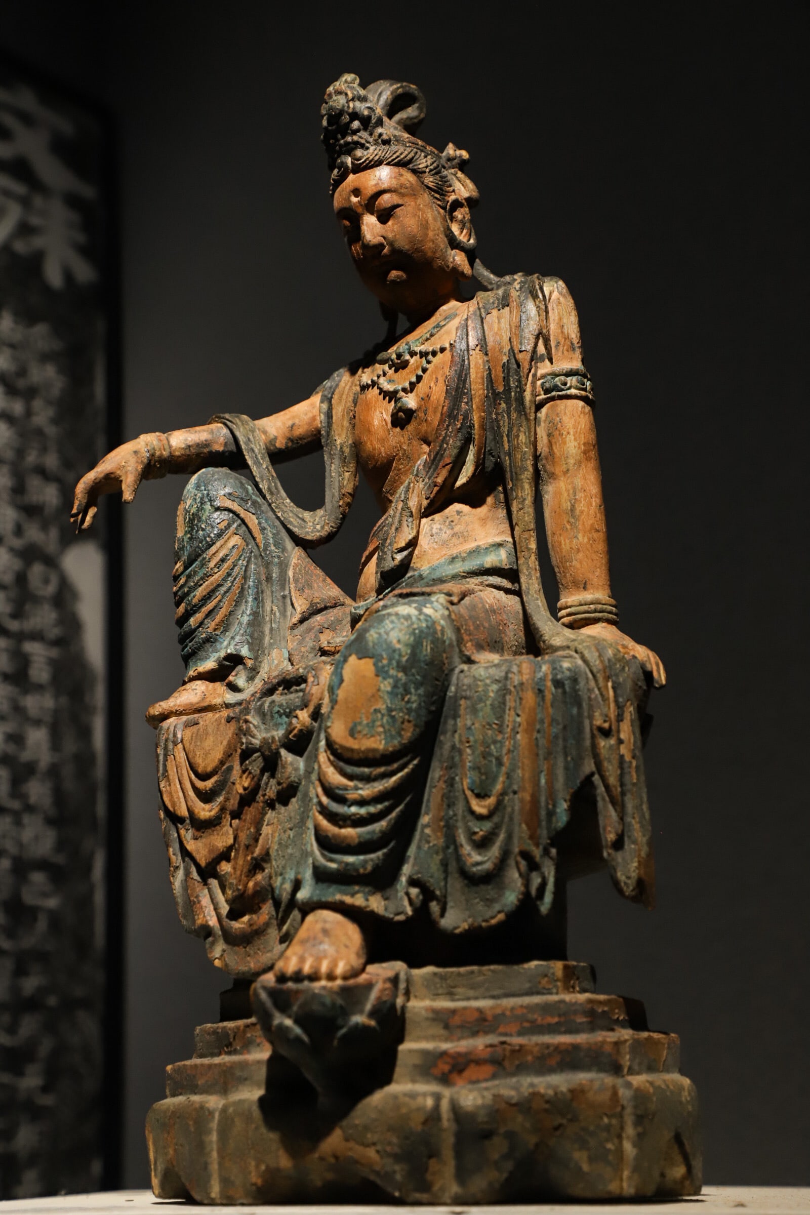 FREE-SPIRITED GUANYIN IN PAINTED WOODCARVING FROM THE MING DYNASTY, SITTING STATUE: Carved from single wood block, with elaborate high hair bun, gentle tranquil face and pearl necklaces on chest. In relaxed rajalilasana pose, one knee raised, one foot resting on small lotus bud. Fade