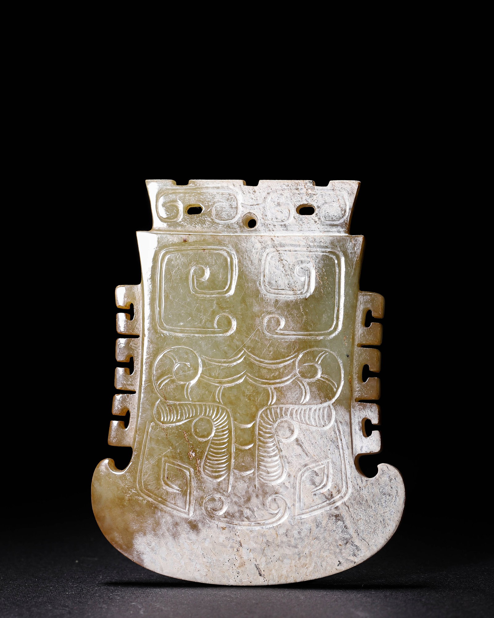 Hetian Celadon Jade Axe-Shaped Pendant with Beast-Mask Pattern, Western Zhou (1046-771 BC): Carved from Hetian celadon jade with partial pale calcification. Formed as an axe-shaped pendant with notched edges and suspension holes. The surface is incised with classic Western Zhou beast-mask mo