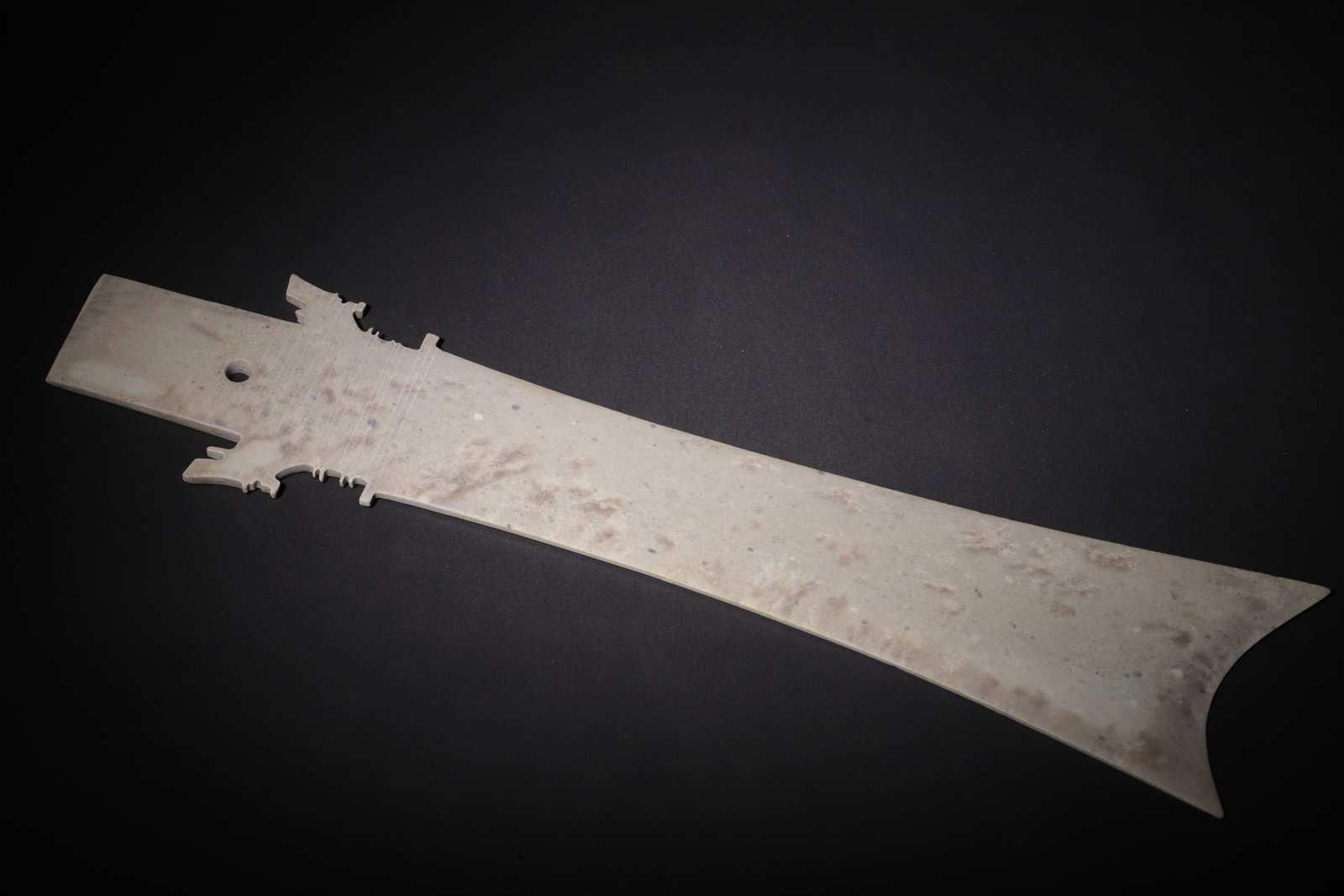 Archaic Jade Zhang Blade, Shang Dynasty (C.1600-1046 BC): The jade is largely calcified with pale brown burial mottling. Elongated flat form terminating in a bifurcated curved blade. Symmetric elaborate serrated flanges extend from both sides of the tang, wh
