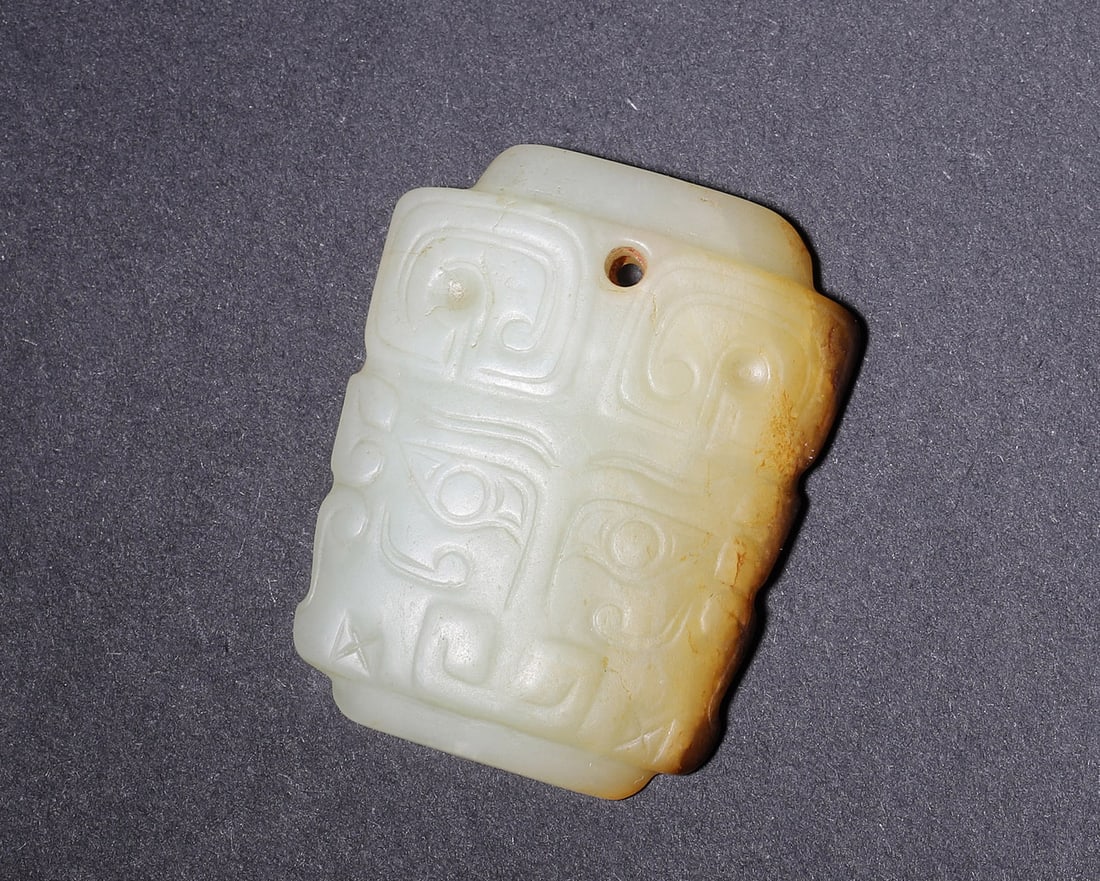 Late Shang Dynasty, White Hetian Jade Beast Mask Pendant with Russet Inclusions: Flat trapezoidal pendant carved from white Hetian jade, single suspension hole at upper edge. Obverse carved with symmetrical double beast masks in shallow relief with classic Shang cloud scrolls and 