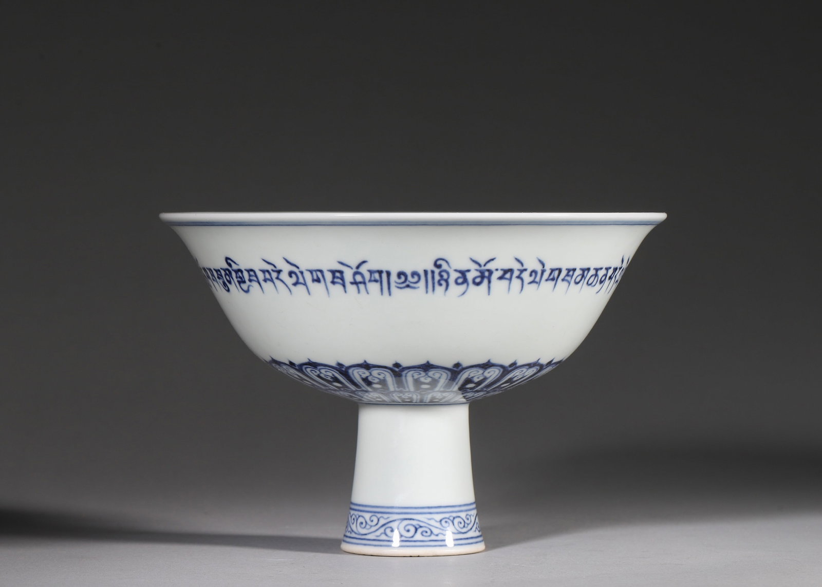 Ming Dynasty, Blue and White Tibetan Script Stem Bowl, Xuande Period: Ming Xuande period stem bowl, flared rim, tall stem foot. Exterior painted with Tibetan auspicious mantra in underglaze blue, layered lotus petals near foot, scrolls on stem. Interior pale celadon