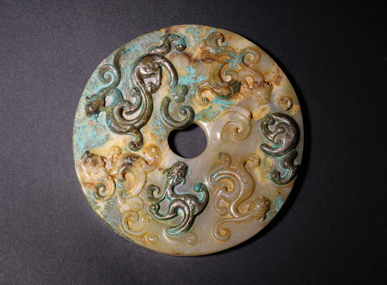 White Jade Bi with High-Relief Chilong and Inscription, Han Dynasty, C.206 BC-220 AD: One side carved with multiple writhing high-relief chilong dragons, reverse incised seal-script inscriptions. Patinated with bronze-green and tan encrustations, a fine Han ritual jade. Dimensions: Dia