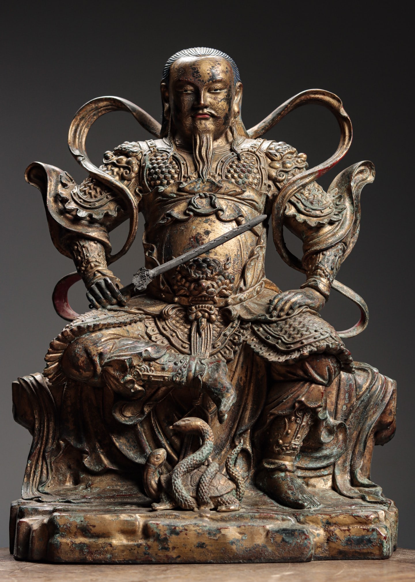 Ming Dynasty, Gilt-Bronze Seated Figure of Zhenwu Emperor: With a round, full face, large pendulous ears, and half-closed eyes, he has loose hair and bare feet. His left hand rests on his knee, right hand holds a sword. Clad in armor, a dragon robe, and a sas