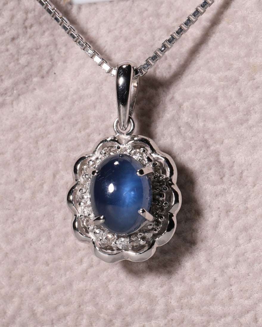 An 18K White Gold Oval Sapphire Pendant Necklace with Halo Diamond Setting: Vertical Drop Set with cabochon sapphire in rich royal blue, this pendant is framed by scalloped 18K white gold and encircling accent diamonds. Paired with delicate box chain, it features vintage eleg