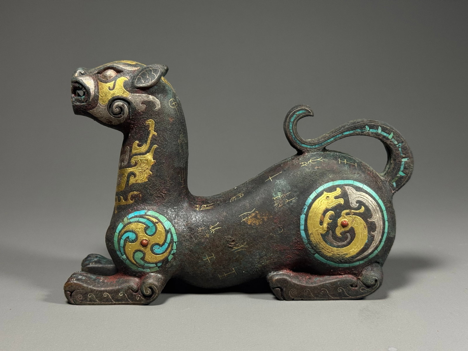 Han Dynasty, Gold, Silver and Turquoise Inlaid Bronze Auspicious-Beast Mat-Weight: Han Dynasty, Bronze Mat-Weight in Form of Auspicious Beast, Inlaid with Gold, Silver and Turquoise.The beast is cast in recumbent posture with raised head and open mouth, body stretched and tail elega