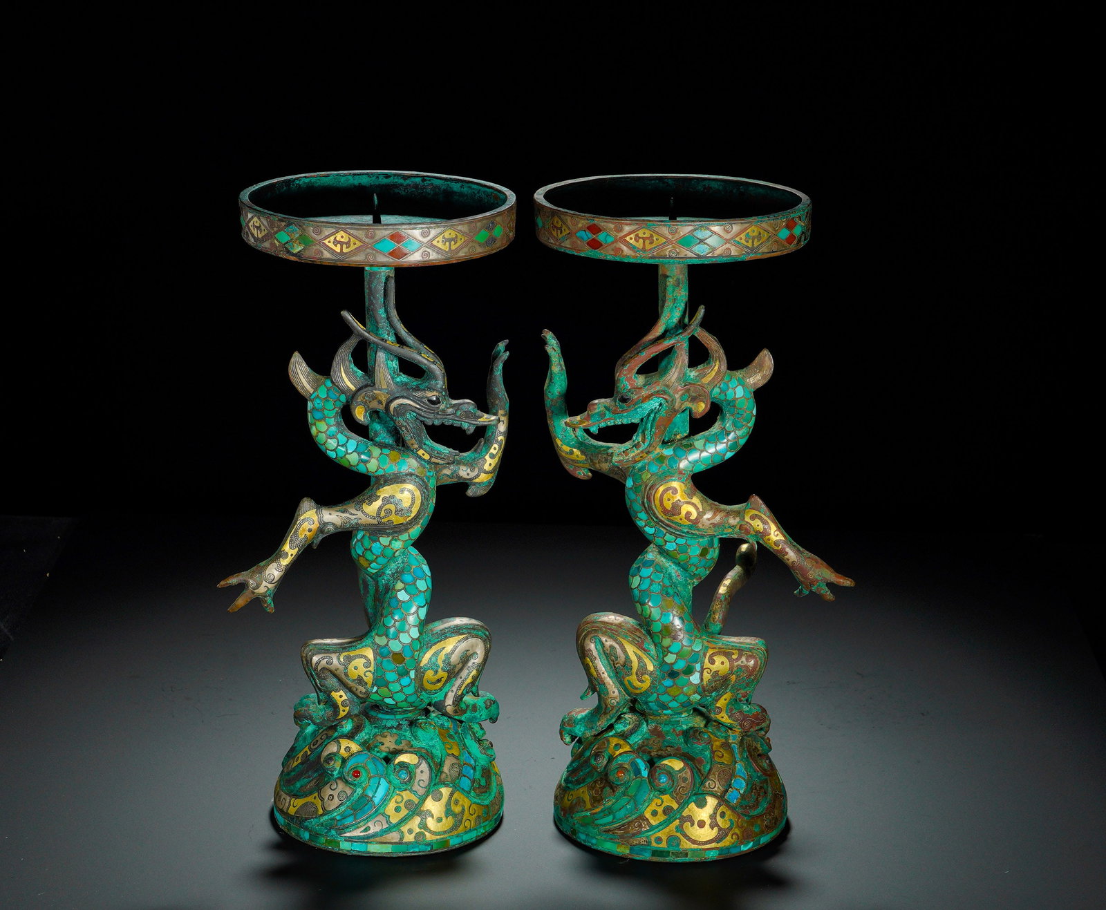 Han Dynasty, Bronze Inlaid Gold and Silver Inlaid Turquoise Dragon-Shaped Lamps (Pair): The pair of Han-dynasty bronze lamps consists of lamp tray, dragon-form stem and base. The shallow open lamp-tray rim is decorated with gold-silver inlaid geometric rhombus patterns. The stem is model