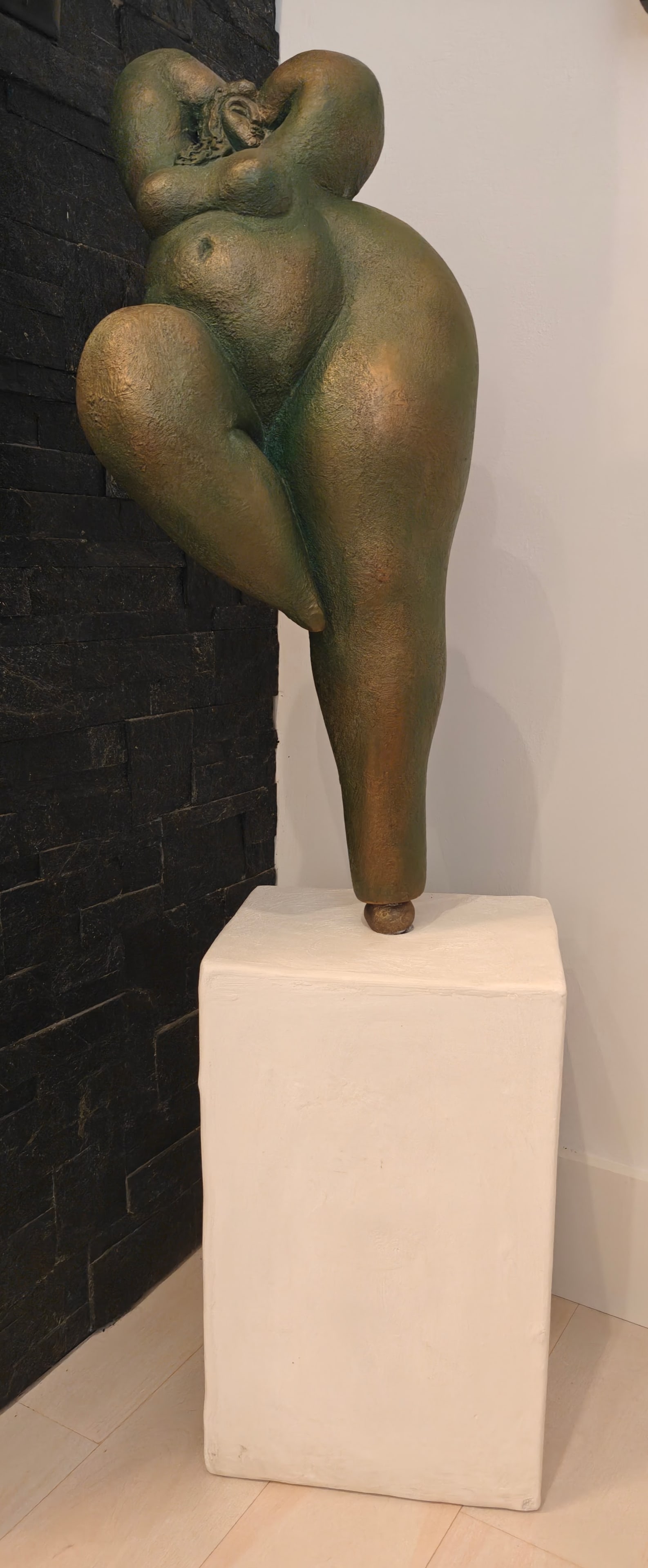 Modern Bronze Female Figure on Base: Large stylized bronze nude female figure with exaggerated voluptuous form, finished in a green and gold patina. Mounted on a white display base. Figure measures approximately 10 x 9 x 24 inches; overa