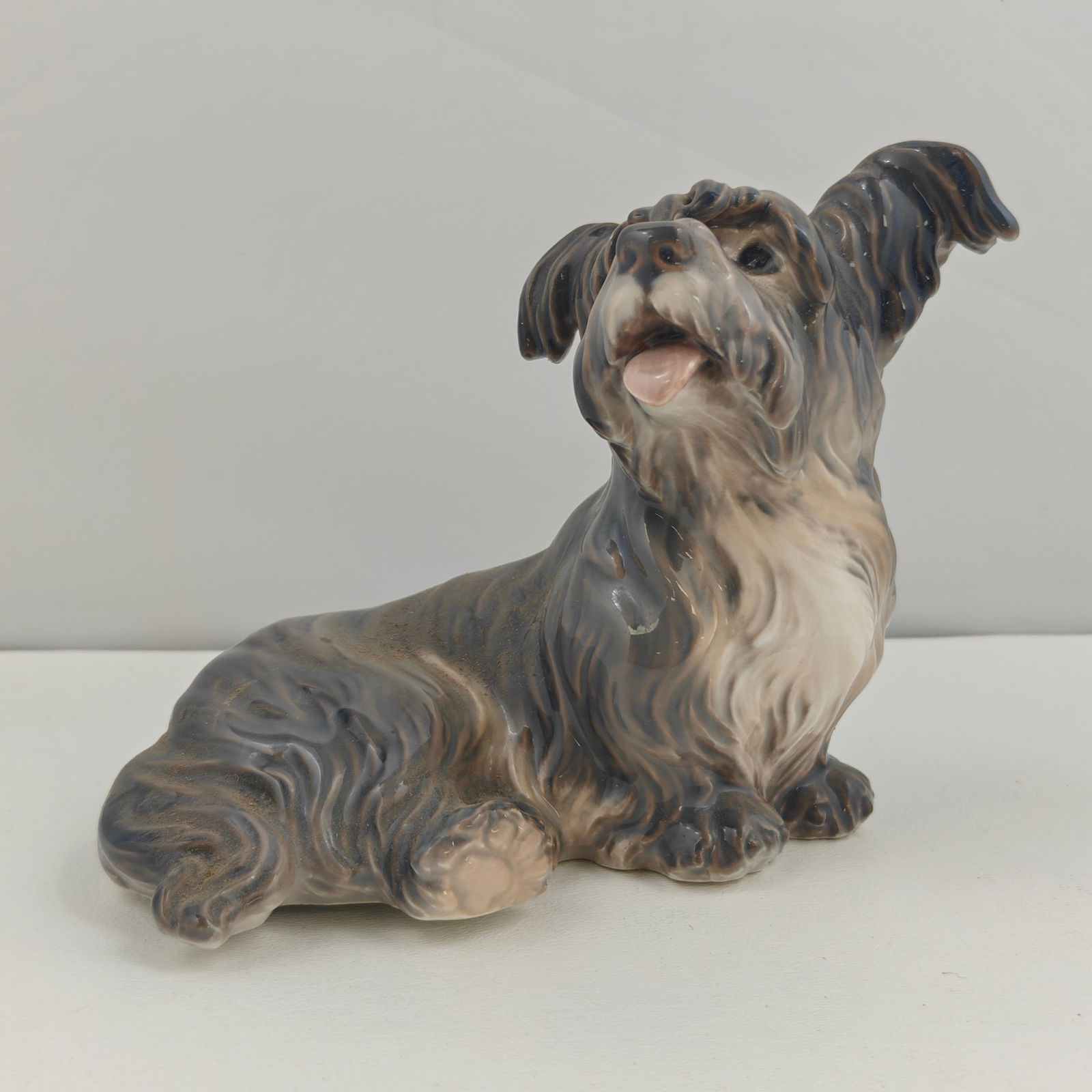 Dahl Jensen Skye Terrier Figurine DJ1103: A Dahl Jensen porcelain figurine of a Skye Terrier, model number DJ1103. Finely hand-painted in shades of gray, black, and tan with lifelike detail, depicting the terrier seated with tongue out and ea
