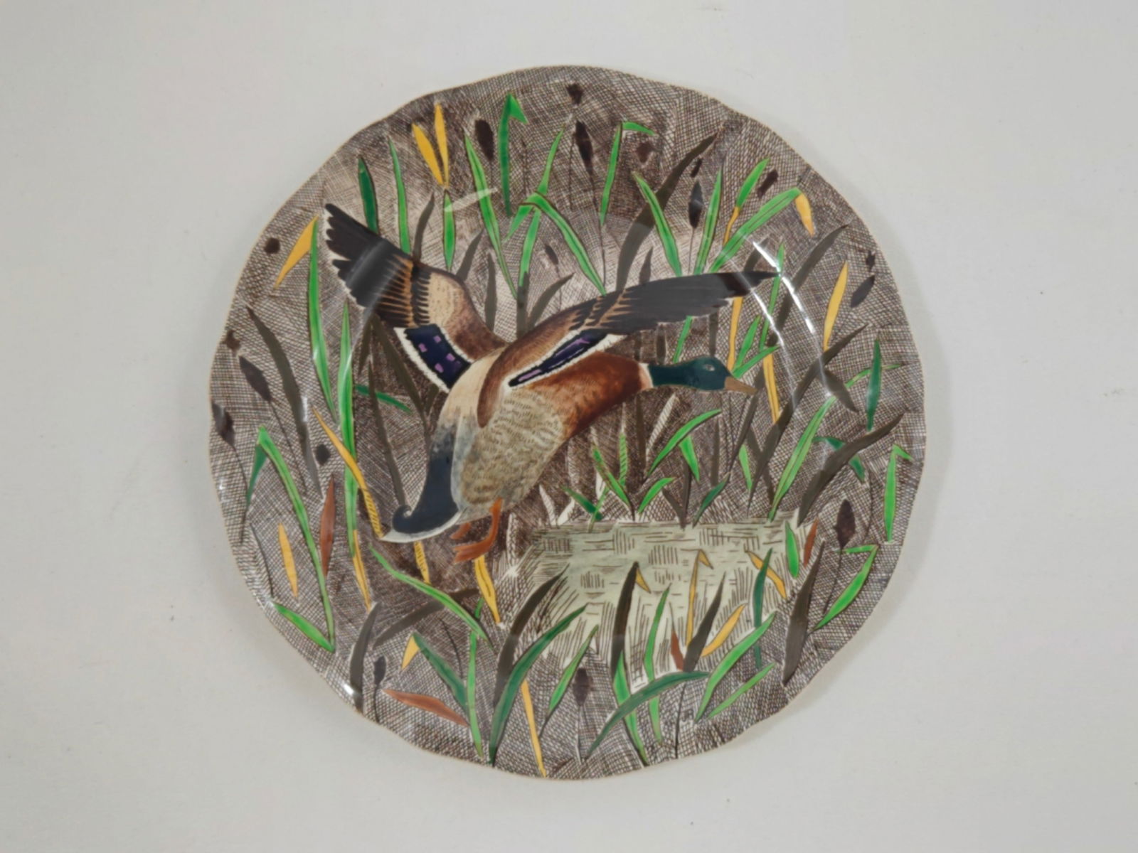 Gien France Faience Dinner Plate with Mallard Duck Decoration: A French faience dinner plate from Gien, decorated with a dynamic scene of a mallard duck in flight amid tall marsh grasses. The composition features earthy brown tones with vibrant green and yellow r