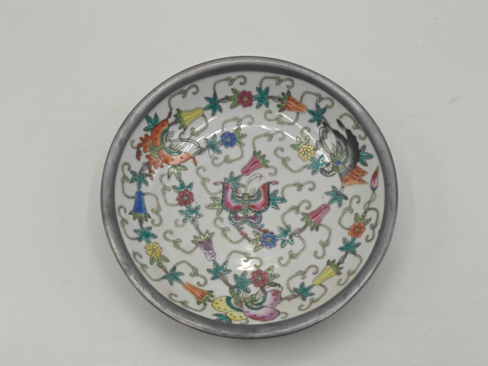 Chinese Porcelain Butterfly Plate with Pewter Mount: Chinese porcelain plate decorated with a central butterfly motif surrounded by scrolling floral and foliate designs in famille rose enamels, set within a fitted pewter surround. Underside bears a red