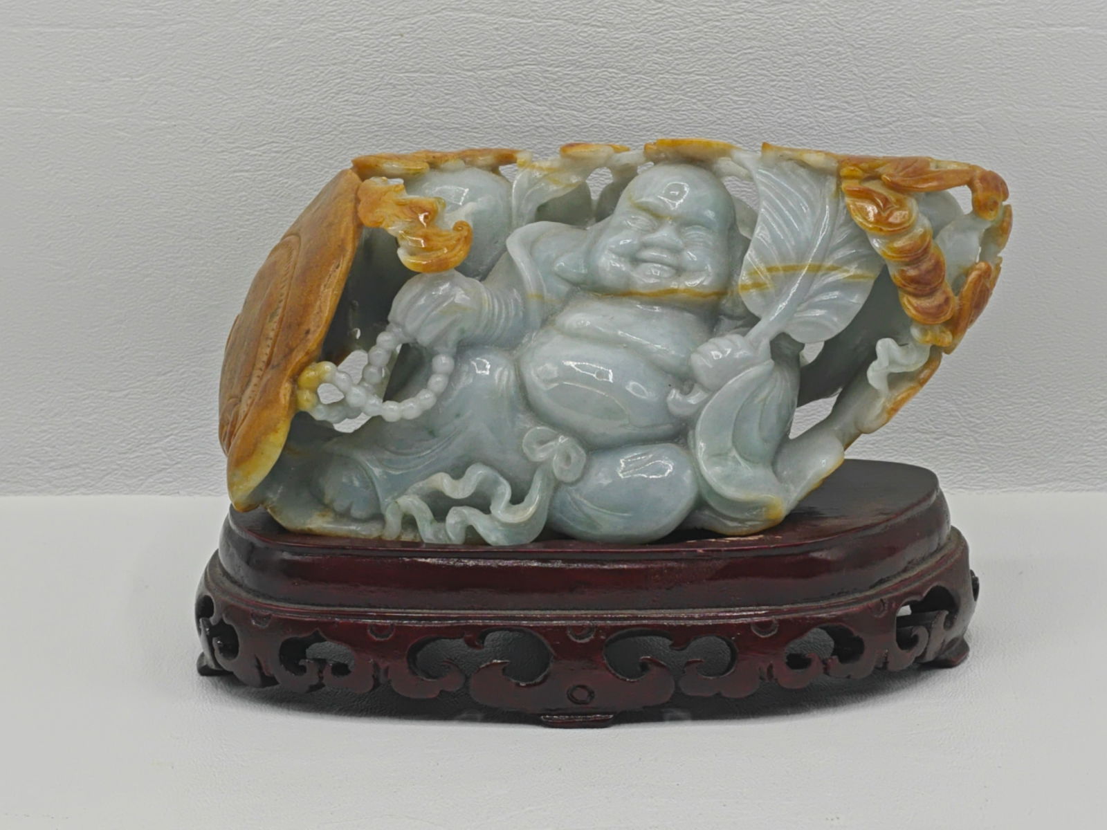 Hand Carved Jade Reclining Buddha Sculpture on Wooden Stand: Hand carved jade sculpture of a reclining Laughing Buddha (Budai), symbolizing happiness, prosperity, and contentment. The figure is finely detailed with flowing robes, prayer beads, and a relaxed, jo