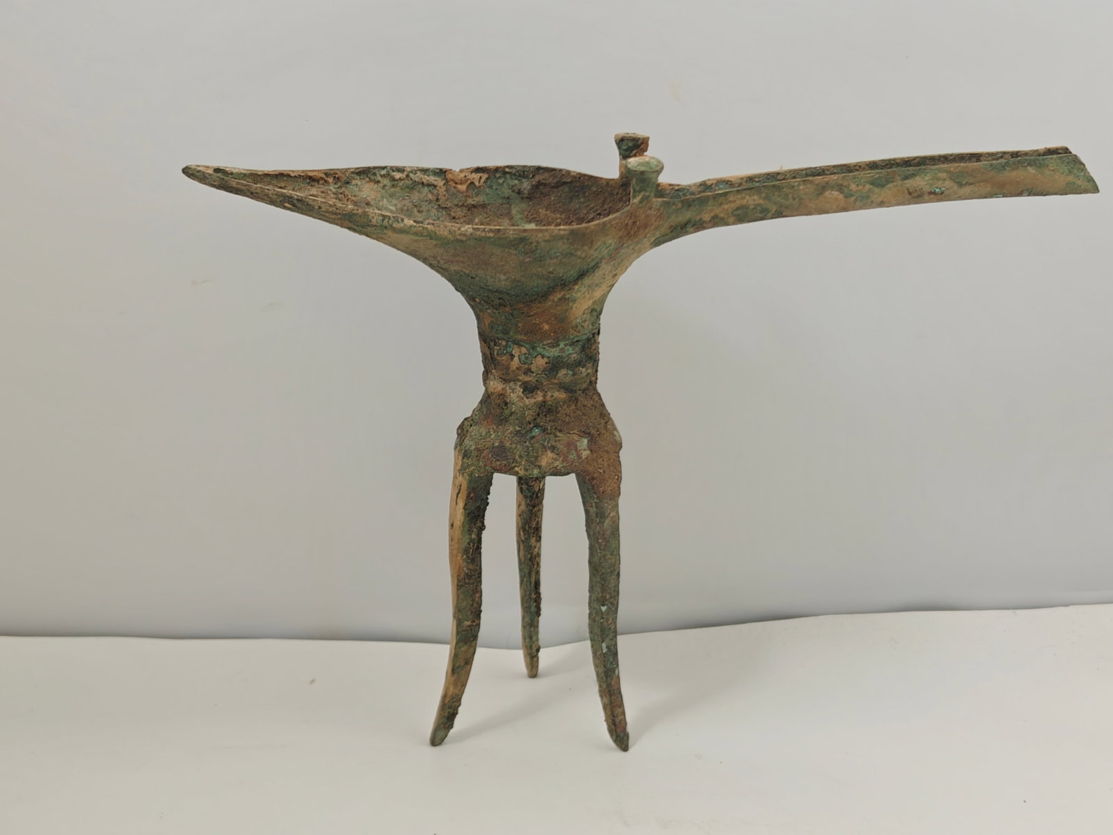 Antique Chinese Bronze Jue Ritual Wine Vessel on Three Legs: Antique Chinese bronze ritual wine vessel, or Jue, featuring the traditional elongated pouring spout, rear tail, twin finial posts at the rim, loop handle, and tripod legs. The vessel displays substan