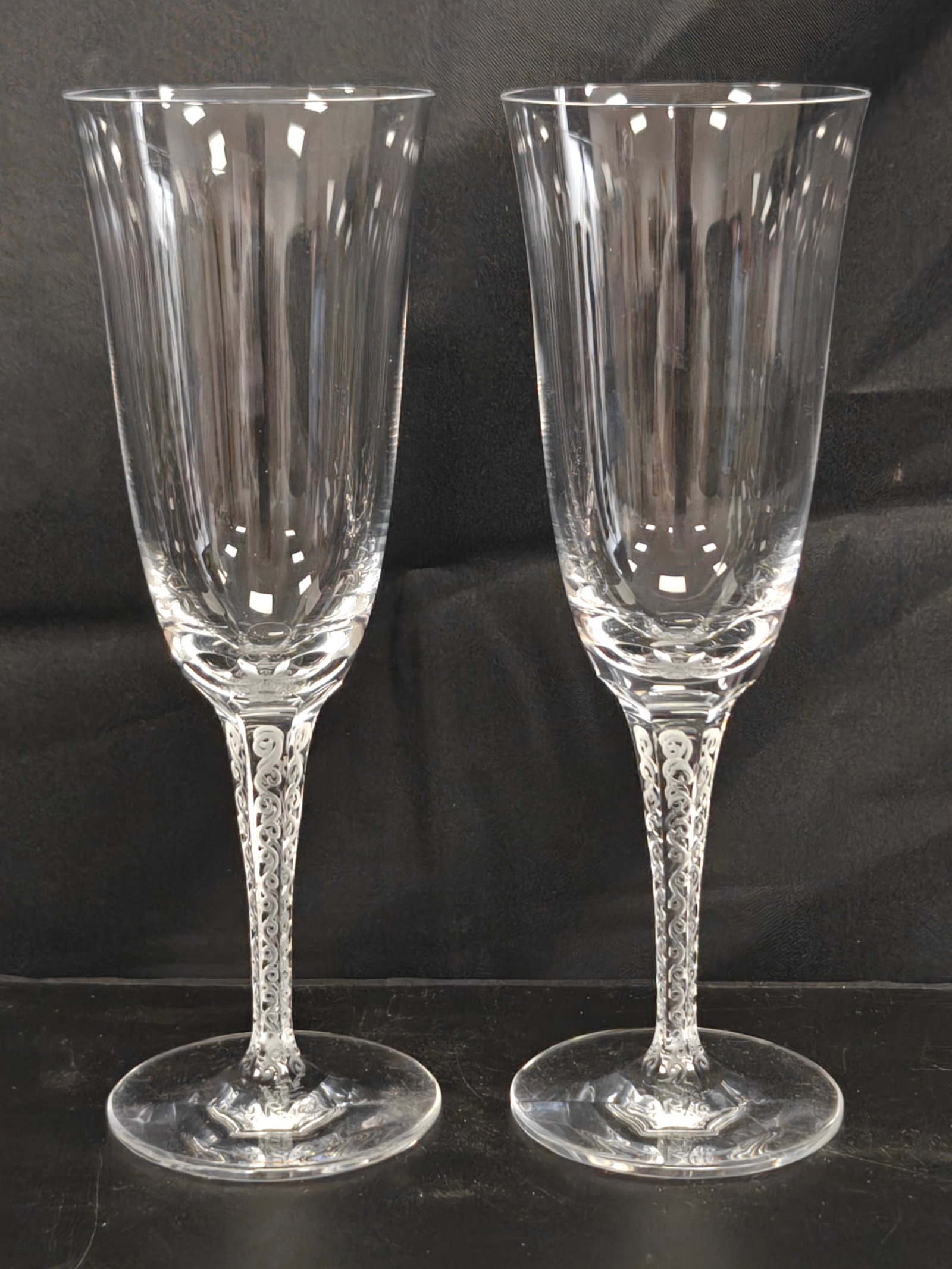 Pair of Lalique France Treves Crystal Champagne Flutes: Pair of Lalique France Treves crystal champagne flutes, each featuring the iconic frosted scroll motif integrated into the hexagonal stem with a clear tapered bowl. Both retain the engraved Lalique Fr