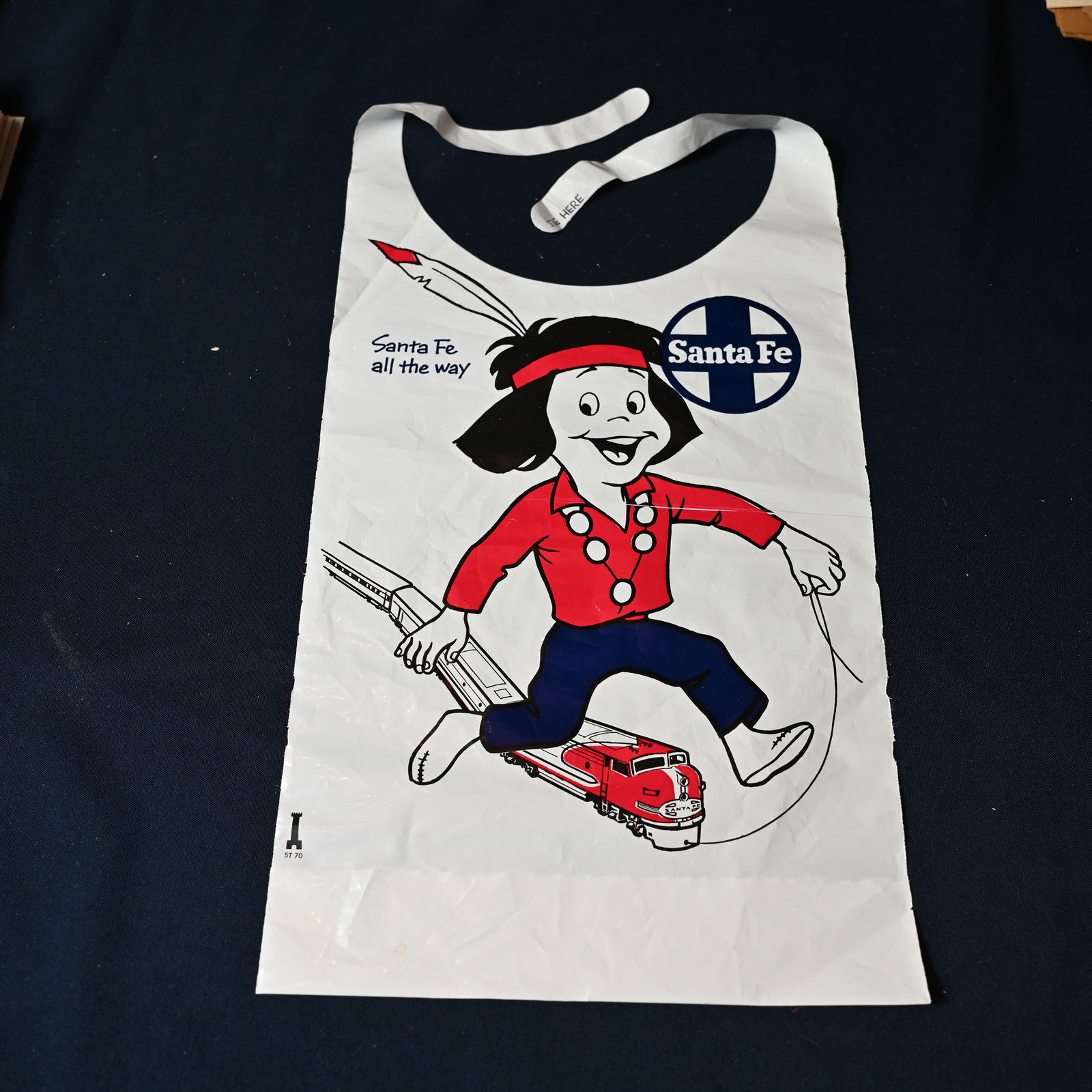 Santa Fe Dining Car Children's Bib: A plastic apron or bib for children with the motto Santa Fe all the way the the company's logo. Measures about 15 inches H ( tip of feather to bottom, excluding tie strips ) by 10 3/4 inches W.