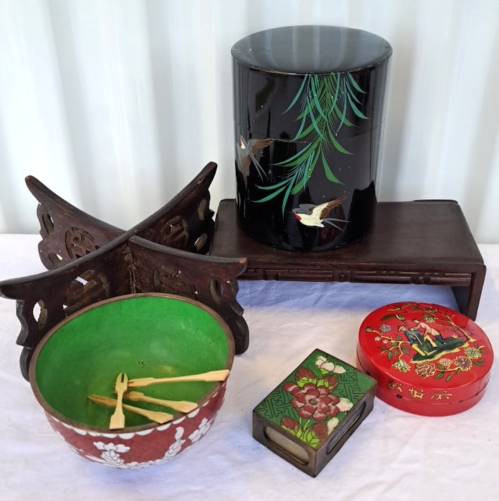 Asian Decorative Lot With Chinese Bowl, Black Tin, Enamel Matchbox Holder Wood Stands: Ornate wood stand has two pieces that slot together. Black tin has scratches. Bowl has a couple of chips inside. Ret tin has a dent on one edge and appears firmly closed. 8 x 8 x 5in. 2lb Low Estimate