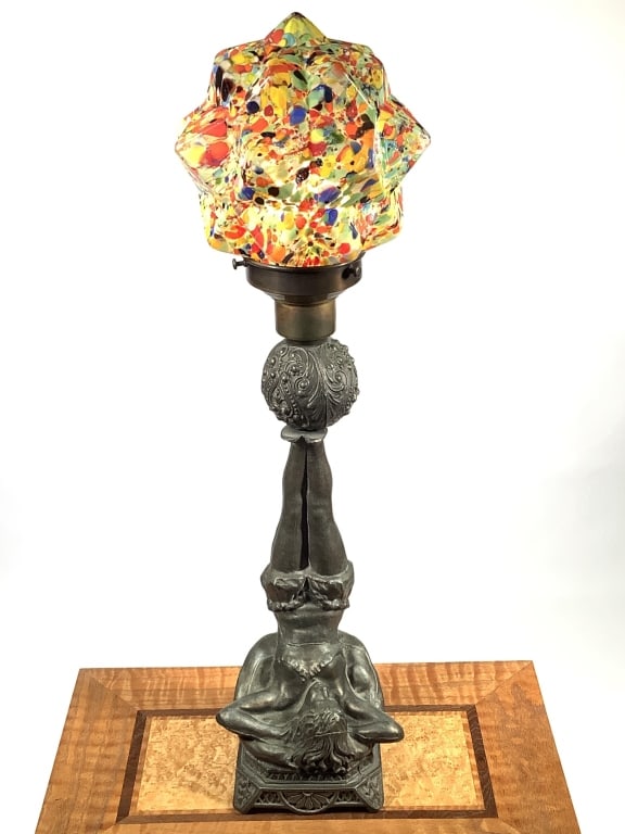 Early 20thC Figural Lamp Base w End of Day Glass: 24"H overall. Figural spelter metal base depicting a young woman, possibly an acrobat, supported by a pillow and with legs raised balancing an ornamented sphere. Geometric form globe in multi-color en