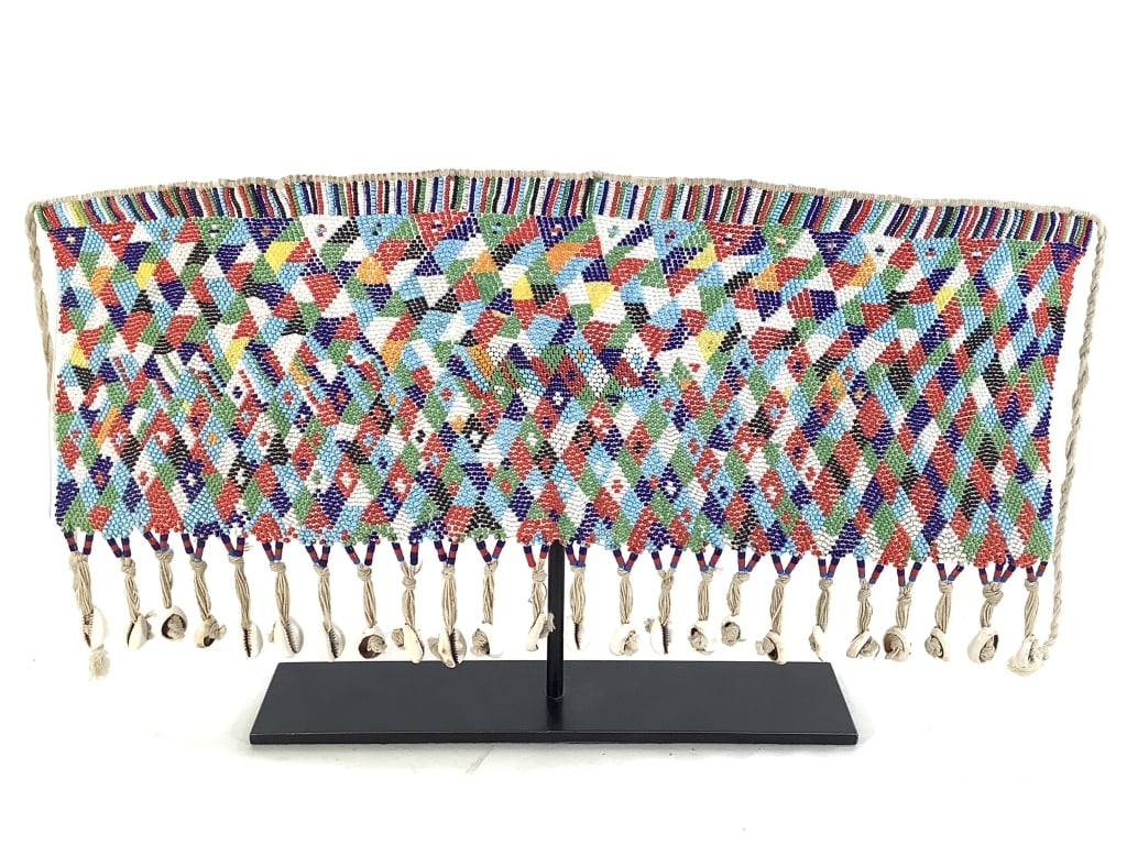 Kirdi Beaded Apron Cameroon: 20 3/4" wide excluding apron strings. Attached to pivoting plexiglas and metal stand. 9 3/8"H on stand. Beaded apron with cowrie shell fringe. Has complex diamond design with an open weave top