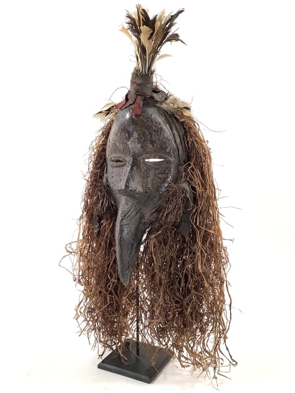 Dan Mask Carved Wood with Raffia West Africa: 22 1/2" H with stand. Carved wood with painted pigment surfaces, raffia, painted and dyed fabric, feathers, metal attached at eyes. Chipping of paint on face. Provenance: Collection of William and