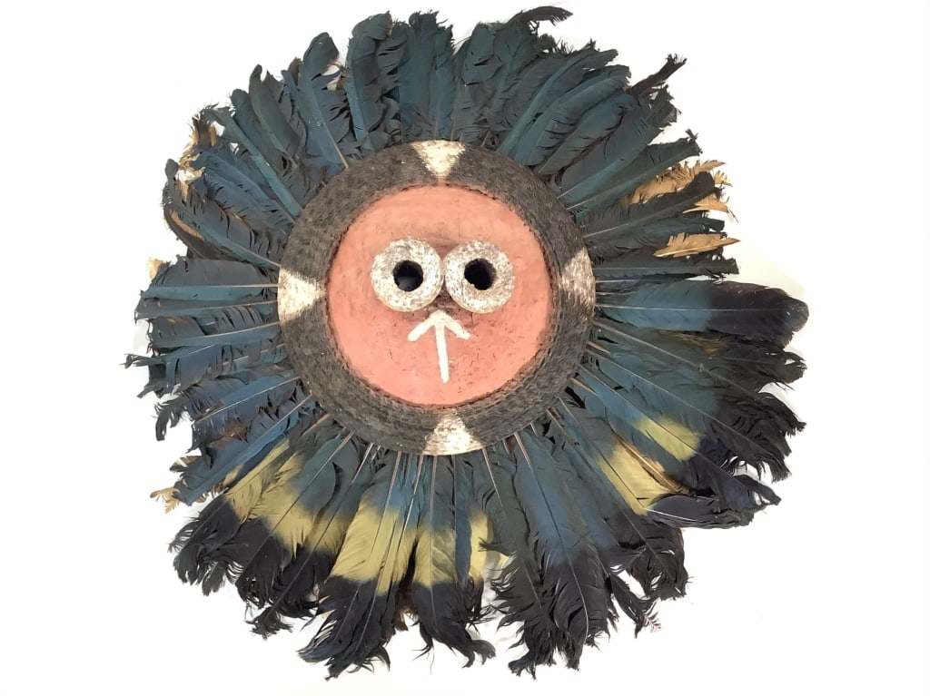 Pende Mask DRC: About 33 1/2" x 33". Woven raffia disk with protruding tubular eyes applied with natural pigment paints and adorned with a ray of Great Blue Turaco feathers. Has a knit raffia head cap on the
