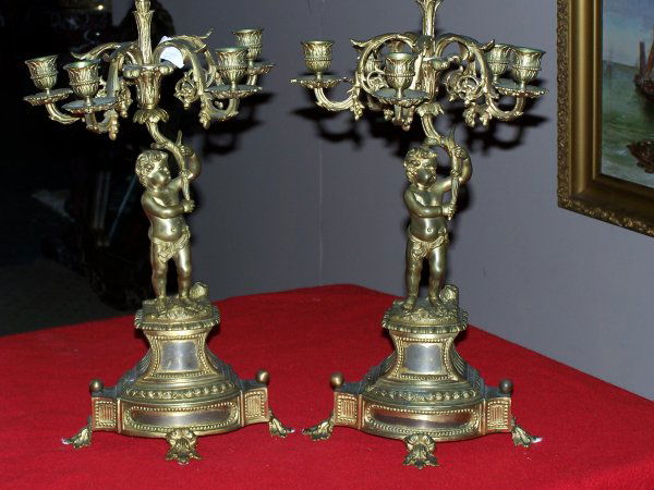 Pair of Brass Candlesticks: Age unknown, but shows wear on the bronze dore. 5 candleholders on each candleabra. Acorn motif in center. Cherubs holding candleabra as center on plinth. 21" H x 10" diameter on leaf feet.