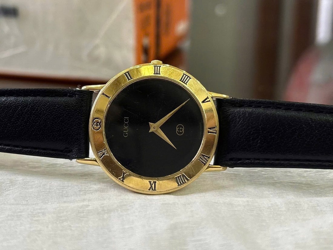 Gucci Swiss Made Gold-Tone Wristwatch: Gucci wristwatch featuring a black minimalist dial with Roman numeral markers and gold-tone hands. Gold-tone case with engraved Gucci logo on the case back. Swiss Made quartz movement. Black leather s