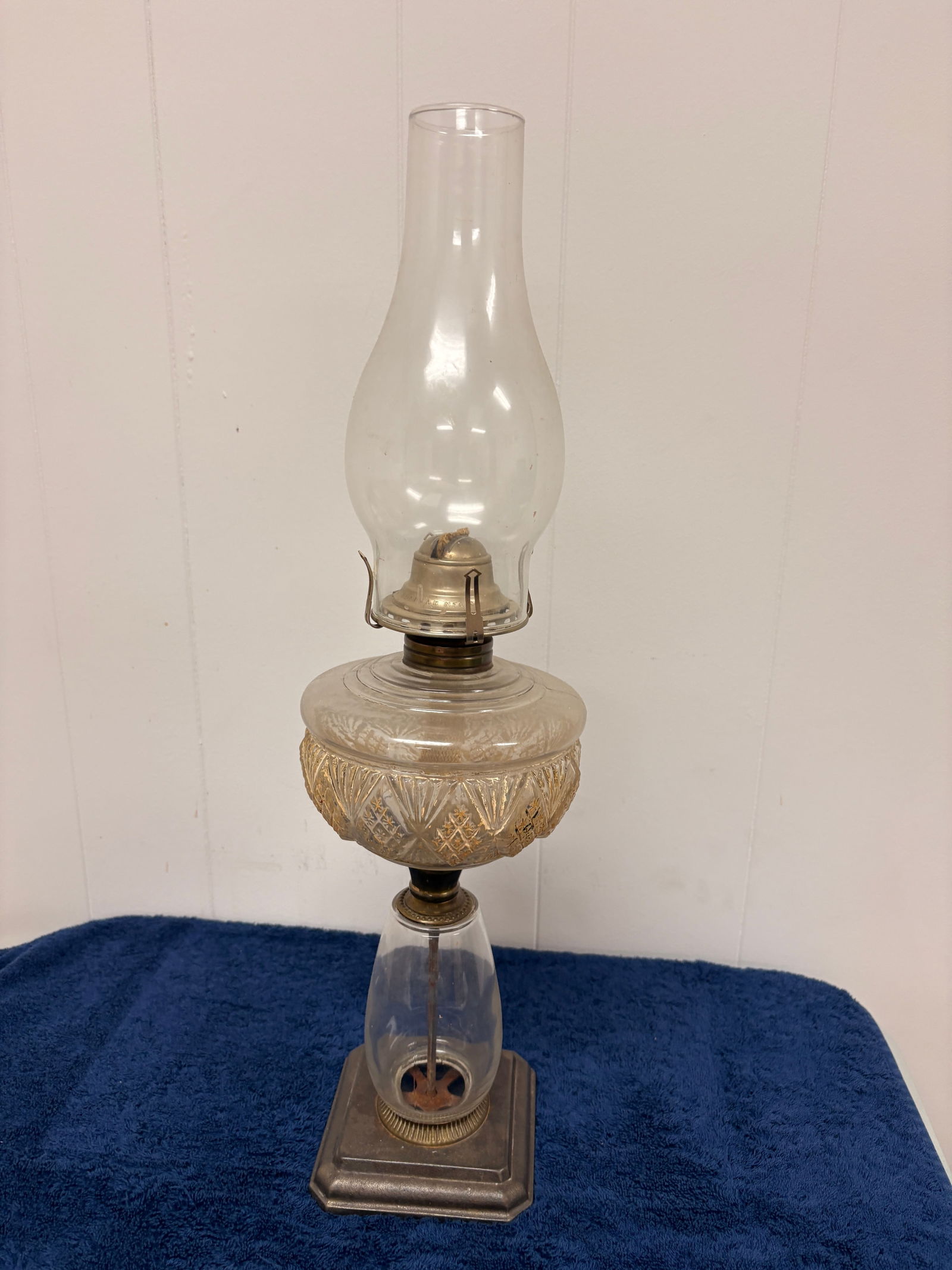Tall Glass Oil Lamp, “Made in Canada” Burner: Tall antique glass oil lamp featuring an embossed glass font, clear glass chimney, and metal burner stamped “Made in Canada.” The lamp stands on a square metal base with visible central wick
