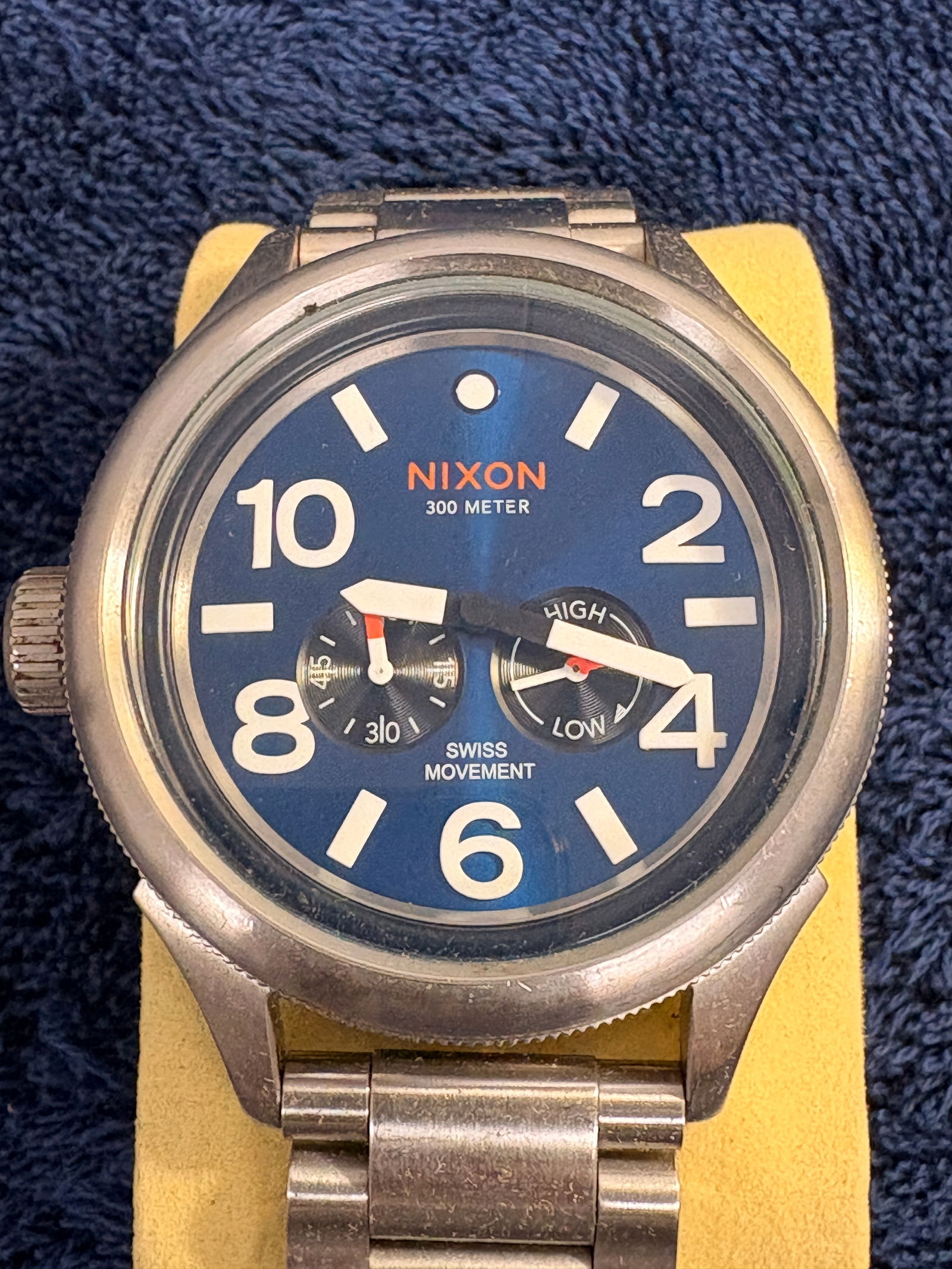 Nixon “The October Tide” Swiss Wristwatch: Nixon men’s wristwatch from The October Tide series featuring a blue dial with large Arabic numerals, dual subdials, and screw-down style crown with orange accent. Brushed stainless steel case and