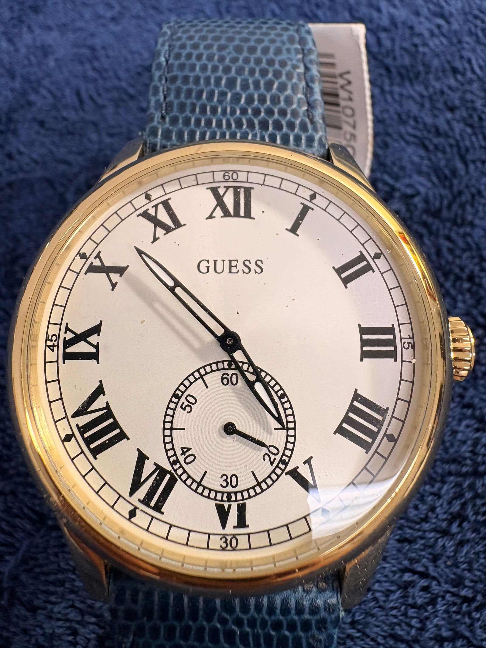 GUESS Classic Roman Numeral Quartz Watch: Elegant GUESS dress watch featuring a gold-tone case with white dial and bold black Roman numerals. Sub-seconds register at 6 o’clock. Fitted with a blue textured genuine leather strap. Quartz movem