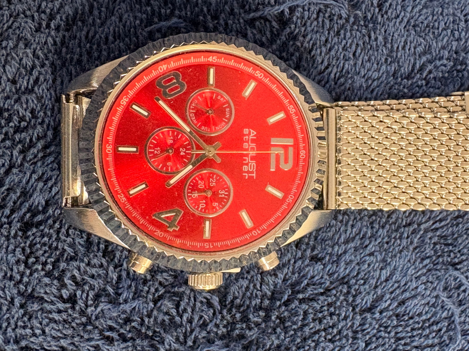 August Steiner Red Dial Multi-Function Quartz Watch: Bold August Steiner wristwatch featuring a striking red multi-function dial with three subdials and textured bezel. Stainless steel case with gold-tone mesh bracelet. Quartz movement. Approx. 46 mm