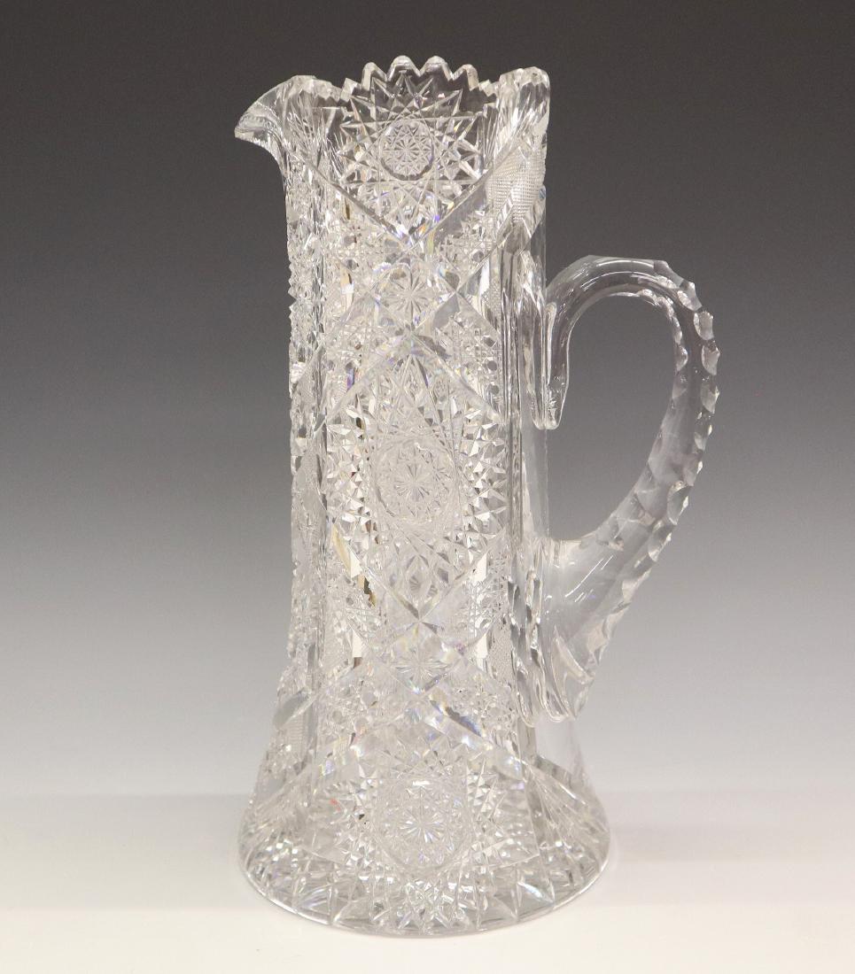 American Cut Glass Pitcher: An American Cut Glass Pitcher. Having a sawtooth rim, star, diamond, cane, and mitre designs. No apparent markings. Small nicks and chips, some scratches at underside. 11 /2" high. ESTIMATE $100-200