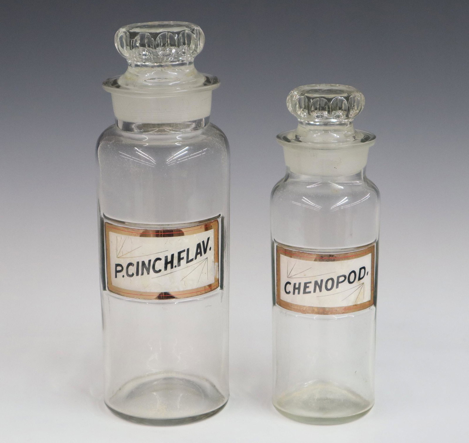 2 Apothecary Bottles: Two turn of the century Apothecary jars. Colorless glass with painted glass labels "Chenopod" and "P.Cinch.Flav" with glass stoppers. Slight wear. Up to 10 1/2" high. ESTIMATE $50-75