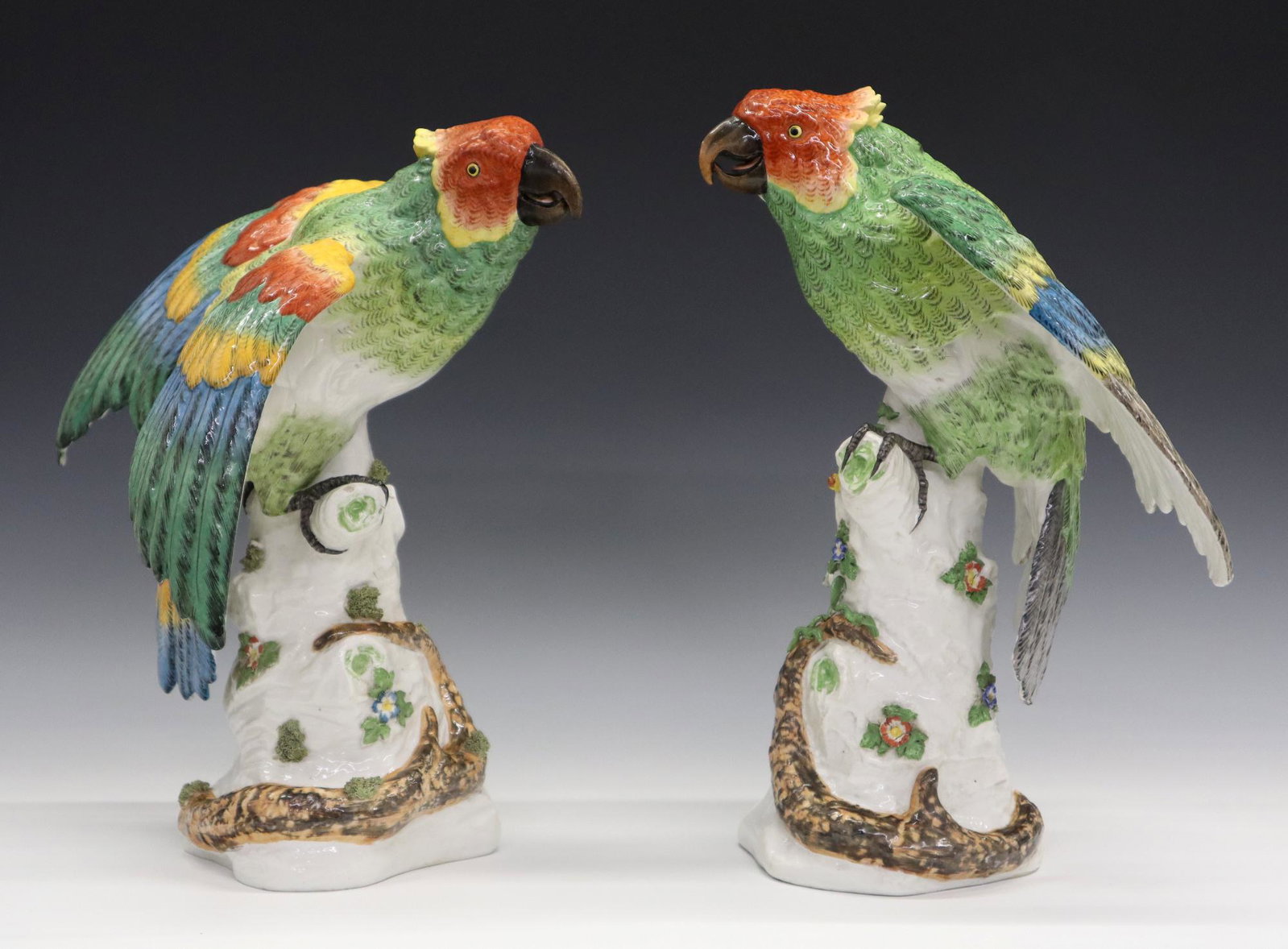French Porcelain Parrots: Two Late 19th Century French Porcelain Parrots. Similarly designed porcelain figures of parrots, both with brightly colored feathers, perched on tree trunks that are clad with flowers, leaves, and vin