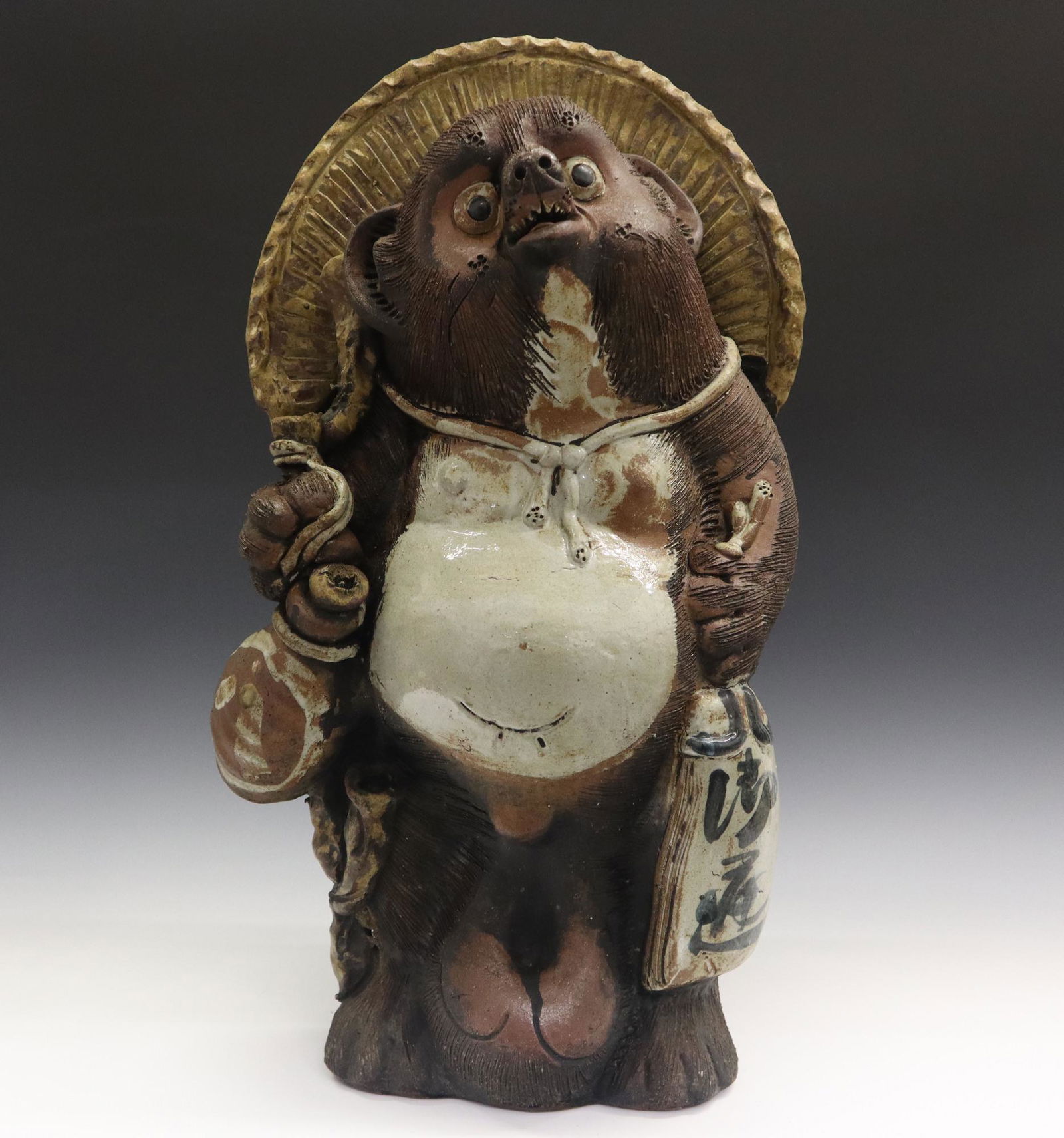 Japanese Tanuki Sculpture: A Japanese Ceramic Sculpture of Tanuki. Glazed ceramic figure of the Folklore version of Tanuki, or a Raccoon Dog, shown wearing a hat and carrying a bottle. Open at the base and at the back of the fi
