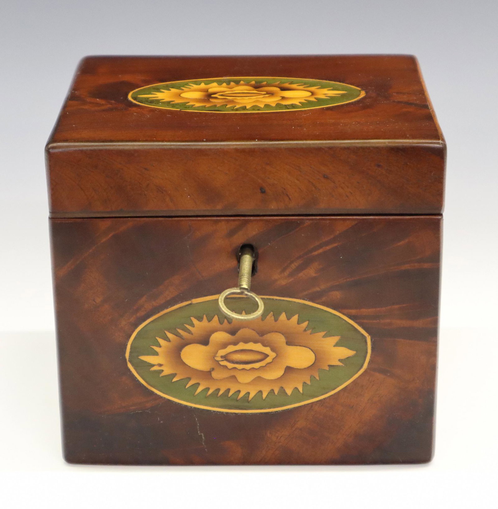 Mahogany Tea Caddy: An early 19th century British tea caddy. Figured Mahogany case with floral inlays on front and lid which opens to reveal a single compartment with foil lining. Refinished, minor wear. 4 1/2" high. EST