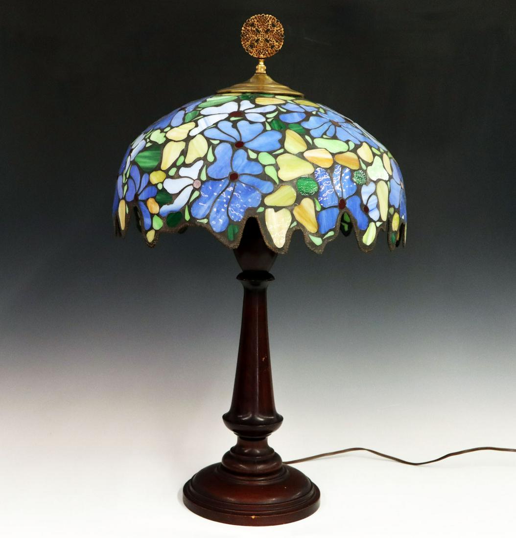 Turned wood lamp base & Repro shade: A Turn of the Century Turned Wood Lamp Base. Mahogany stained base with a Reproduction leaded glass shade with floral motif in blue and green. Wear to base and a 4" check to stem. 16 1/2 x 28" high ov