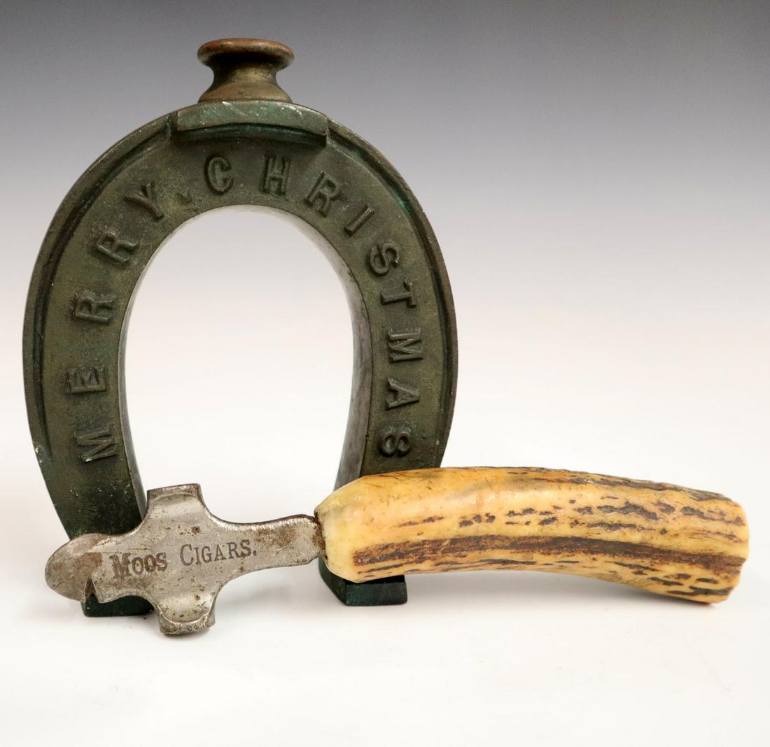 2 Novelty Items: Two Turn of the Century Novelty Items. Including a cigar box opener/hammer with horn handle and "Moos Cigars" advertising logo together with a brass horseshoe paperweight with "Merry Christmas" at one