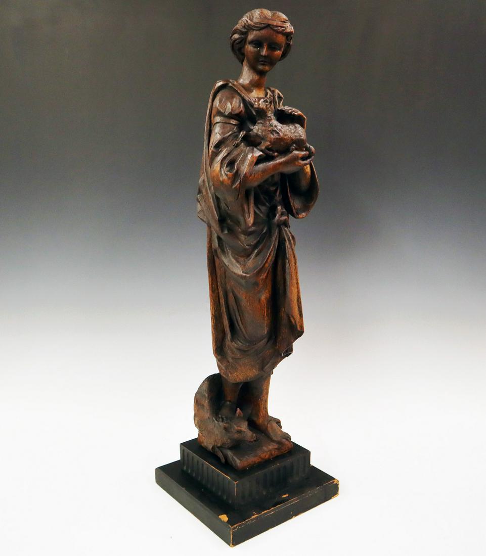 Carved Oak Sculpture of St. Agnes: A Late 19th century Carved Oak Sculpture of St. Agnes. Depicted standing, holding a lamb with a wolf at her feet. Originally a furniture ornament, the base is a later addition. Age cracks to the back 