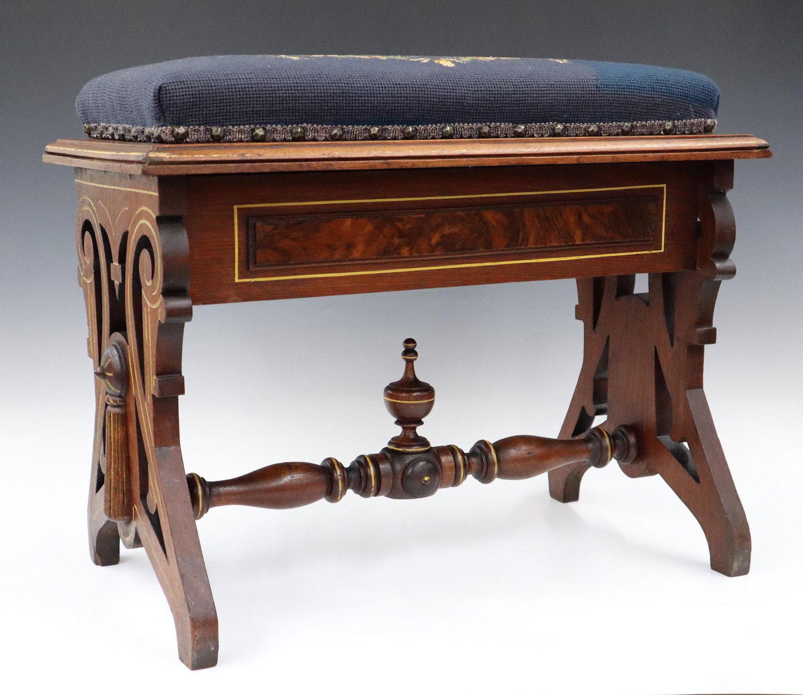 Eastlake Organ Bench: A Late 19th century Eastlake Period organ bench. Walnut construction with gold incised carving, tassles at the ends, turned stretcher, and blue needlepoint seat which lifts for storage. Wear to the go