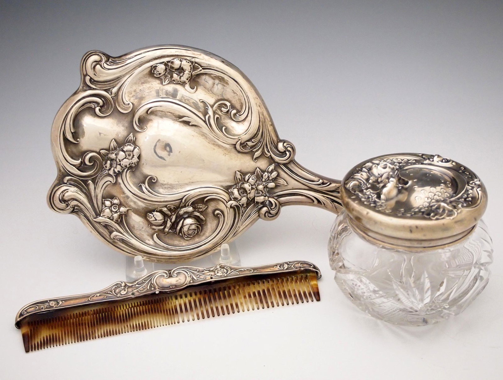 3 Sterling Dressing Table items: Three 20th century dressing table items with Sterling Silver fittings. Includes a 10 1/2" hand mirror, a 7 1/2" comb and a cut crystal jar with Sterling cover. Wear and some denting. ESTIMATE $100-150