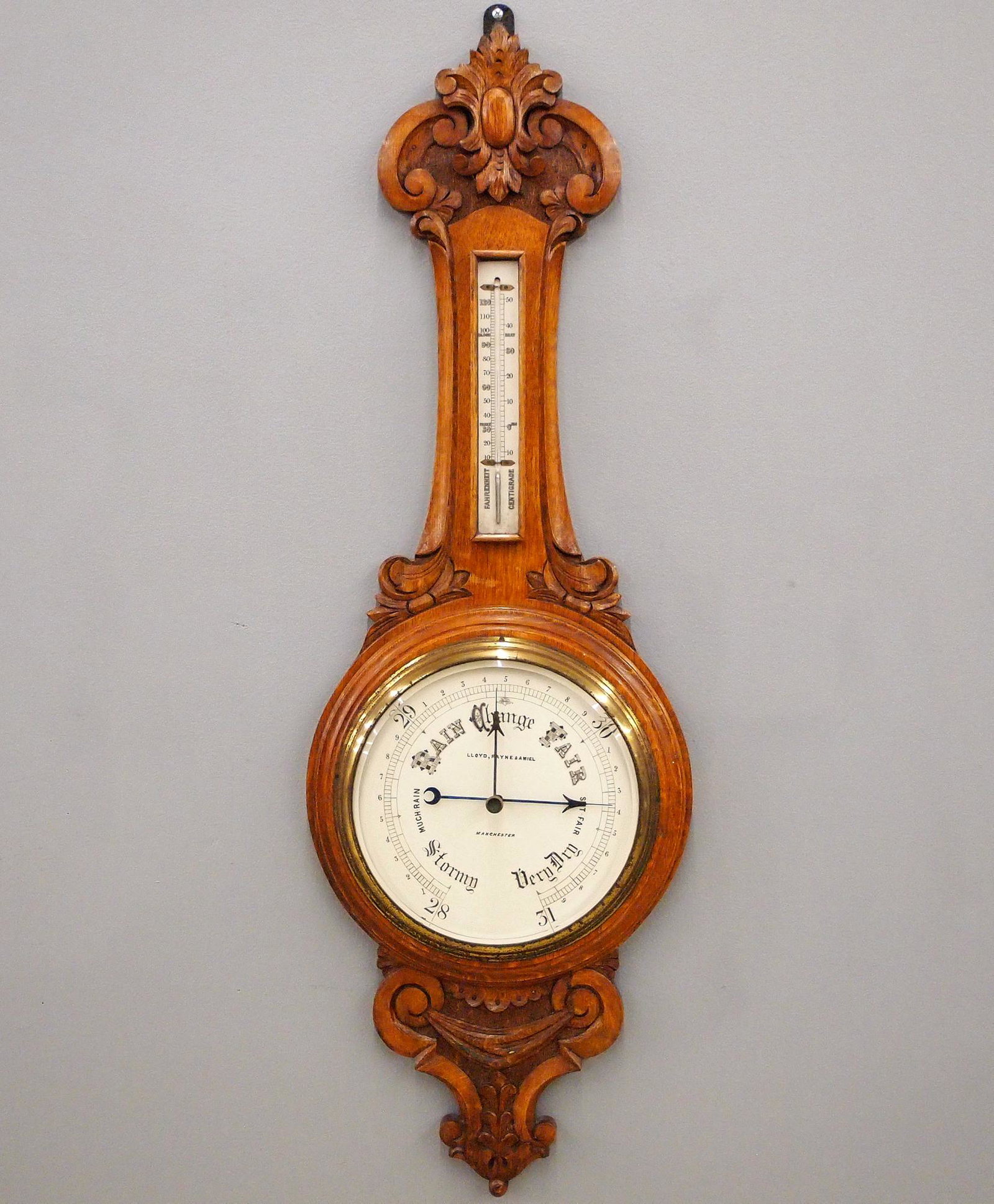 Carved Oak Wall Barometer: A late 19th century English carved Oak aneroid barometer retailed by Lloyd, Payne & Ameil, Manchester. Upper thermometer and lower barometer dial with beveled glass and Brass bezel. Solid Oak case wit