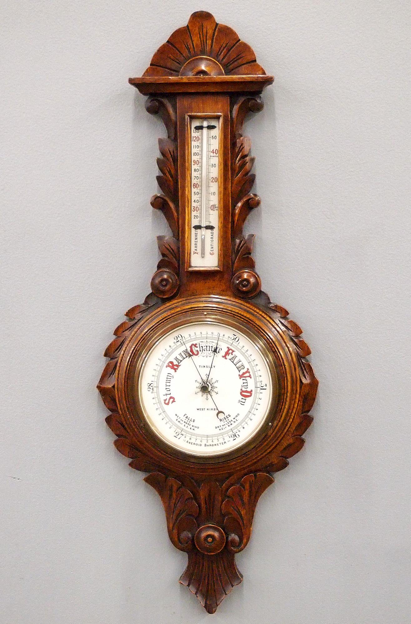 Carved Oak Wall Barometer: A late 19th century English carved Oak aneroid barometer retailed by Tinsley, West Kirby. Upper thermometer and lower barometer dial with beveled glass and Brass bezel. Solid Oak case with carved foli