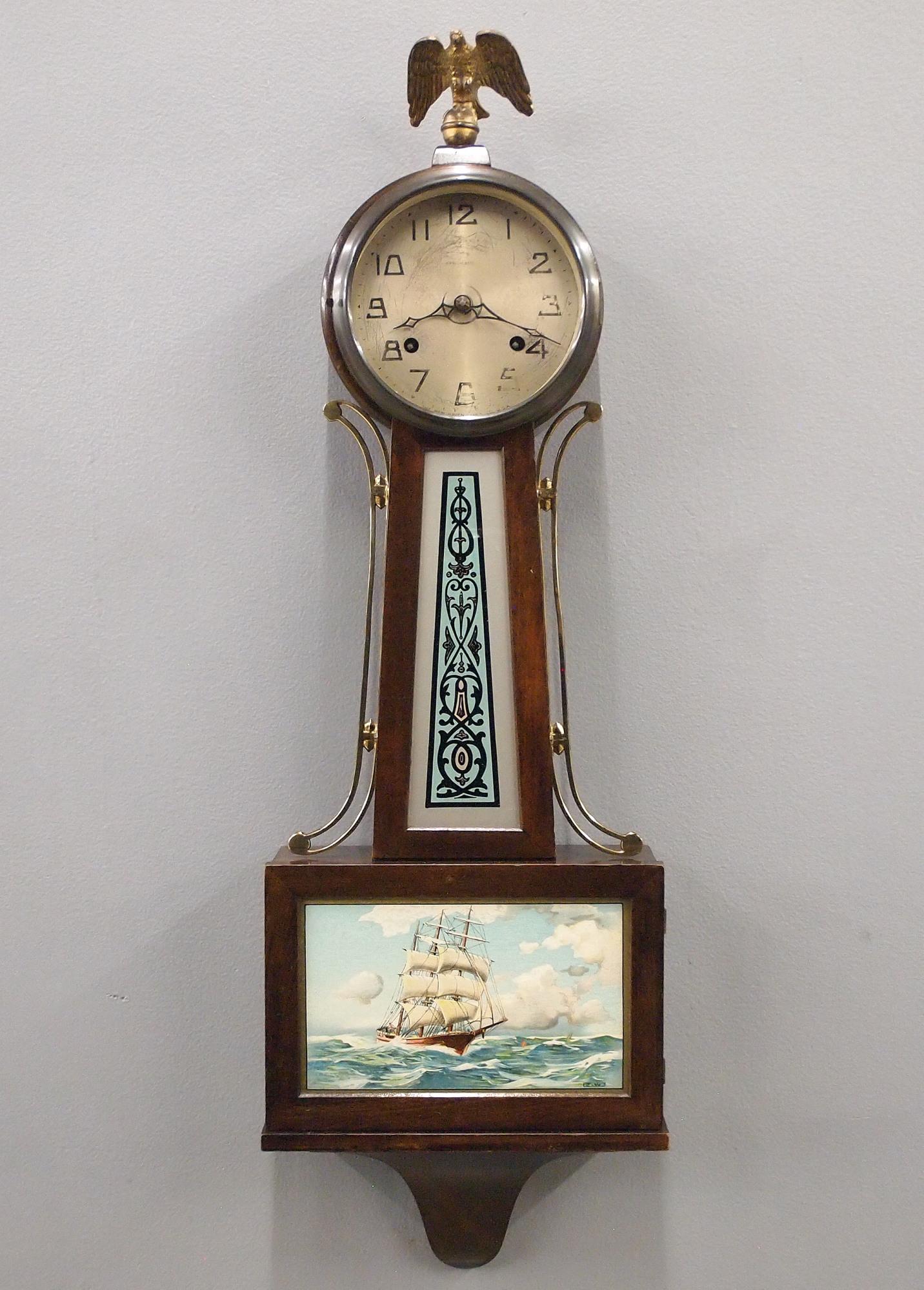New Haven Whitney Banjo Clock: An early 20th century New Haven "Whitney" model Banjo clock. 8-day spring driven time and strike movement with silvered metal dial Mahogany case with Brass arms and Eagle finial, decals on throat and 