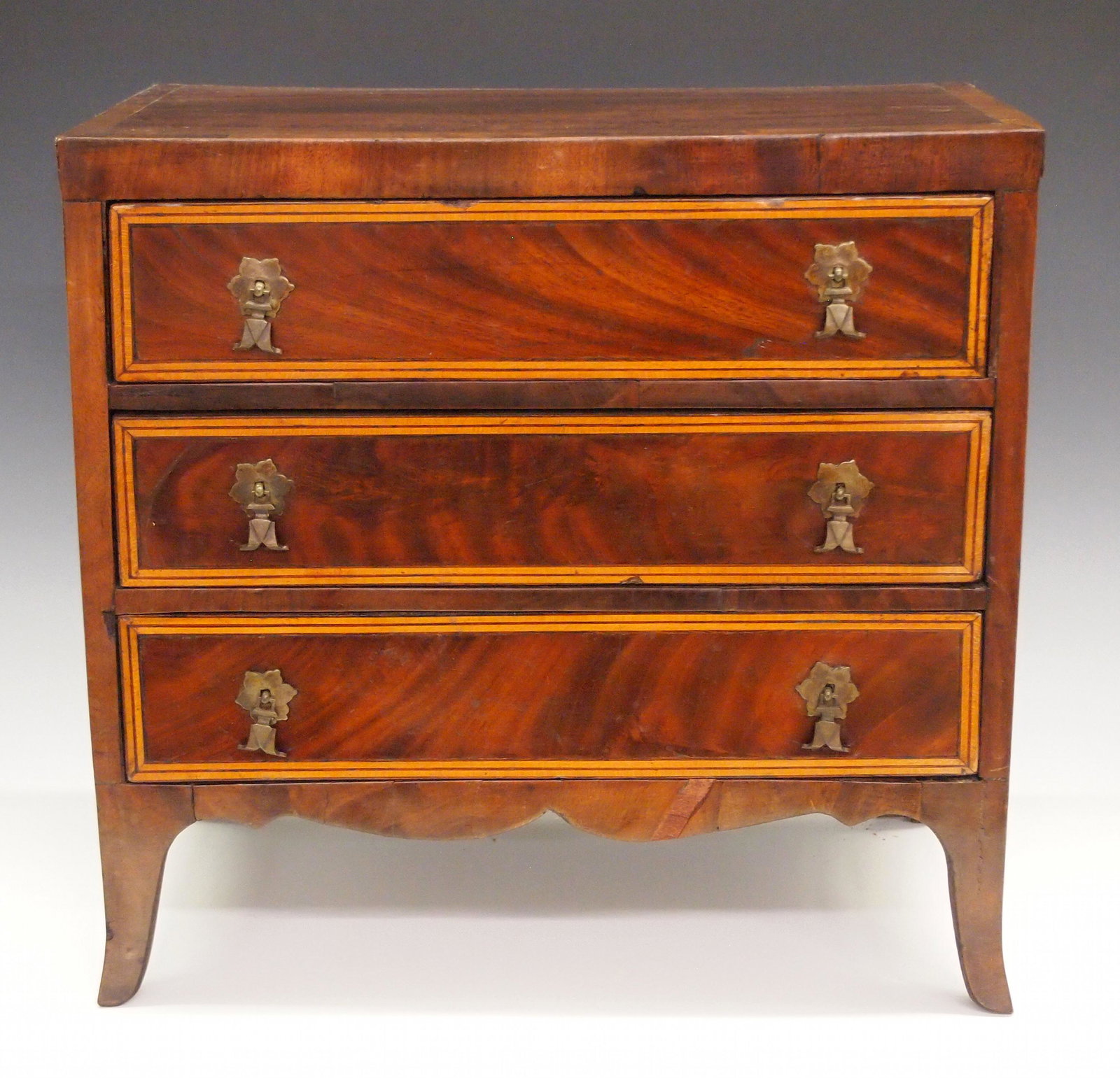 Miniature Hepplewhite Chest: A 20th century miniature Hepplewhite style chest. Mahogany construction with three banded drawers over a shaped apron and tall French feet. Some wear, minor shrinkage and edge damage. 15 1/2 x 9 x 14 