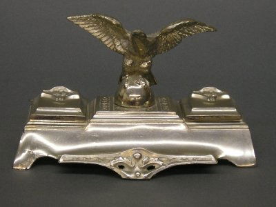 Figural inkwell: An early 20th century figural nickel plated inkwell. An Eagle perched between two inkwells with hinged covers and glass inserts. Wear and denting at foot. Faint makers mark on base. 7 1/2" long. ESTIM